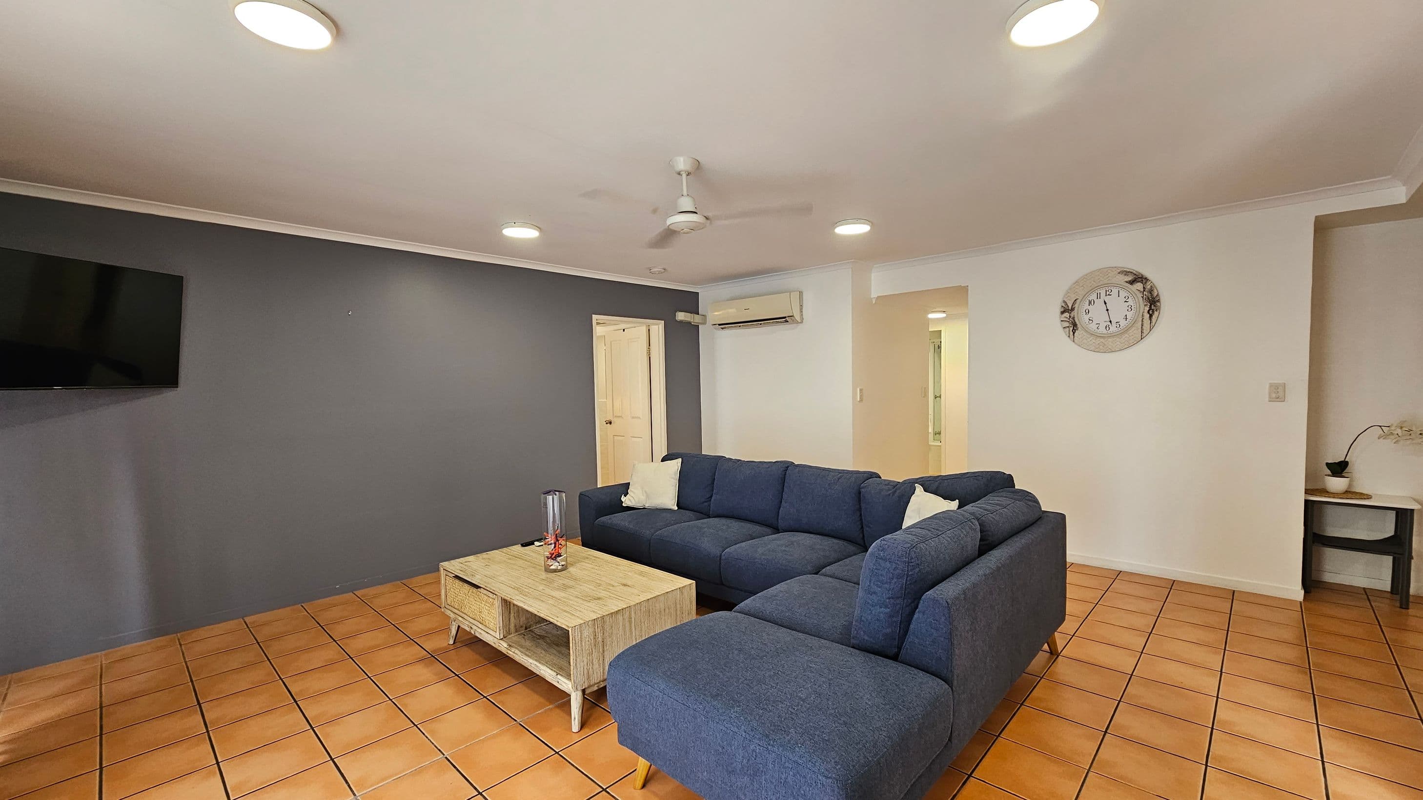 A living room with terracotta tiled flooring and a white ceiling featuring recessed lights and a ceiling fan. On the left, a dark blue accent wall holds a mounted flat-screen TV. In the centre, a large blue L-shaped sofa with white cushions faces a wooden coffee table. On the right, a white wall displays a decorative clock. Below the clock, a small table with a potted plant is visible. An air conditioning unit is mounted above a doorway in the background.