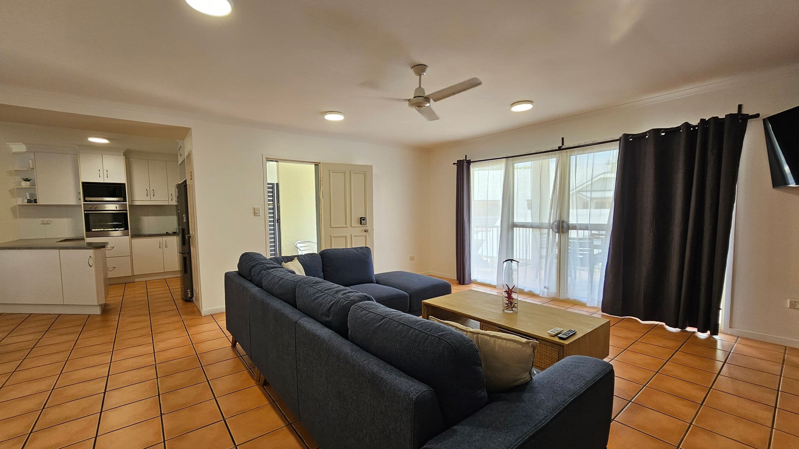 A living room with terracotta tiled flooring and a white ceiling with recessed lights. On the right, a large window with sheer and dark curtains lets in natural light. A dark blue L-shaped sofa faces a wooden coffee table with a decorative item. A flat-screen TV is mounted on the wall. On the left, an open kitchen area features white cabinets, a built-in oven, and a fridge. A ceiling fan is centred above the room.