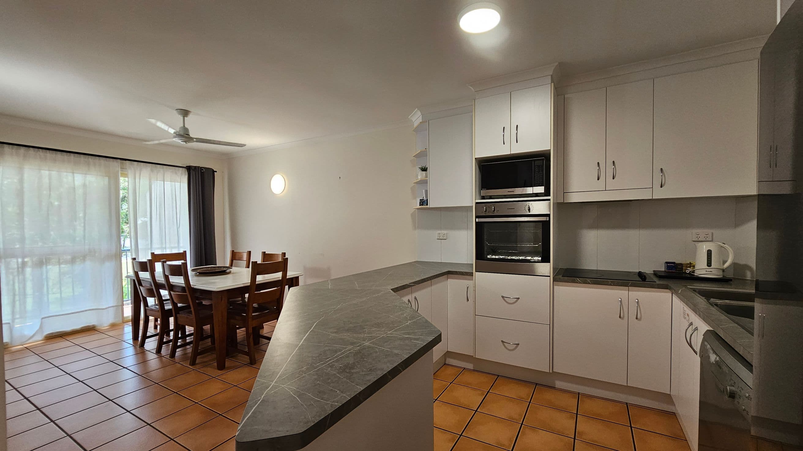 Kitchen and dining area with terracotta floor tiles. On the right, there are white cabinets, a built-in oven, and a kettle on the counter. The kitchen has a dark countertop with a corner sink. On the left, a wooden dining table with six chairs is near large windows covered with sheer curtains. A ceiling fan is above the dining area, and a round wall light is on the wall.