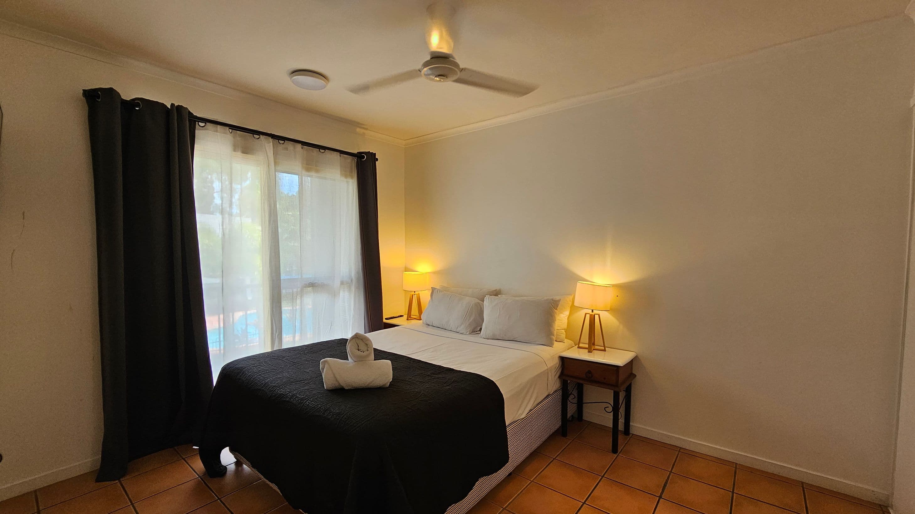 A bedroom with a double bed in the centre, covered with a black quilt and white sheets. Two white pillows are placed at the head of the bed, with a rolled towel on top. On either side of the bed are wooden bedside tables, each with a lit lamp. A ceiling fan is above the bed. To the left, there is a window with sheer white curtains and dark drapes. The floor is tiled in a terracotta colour.