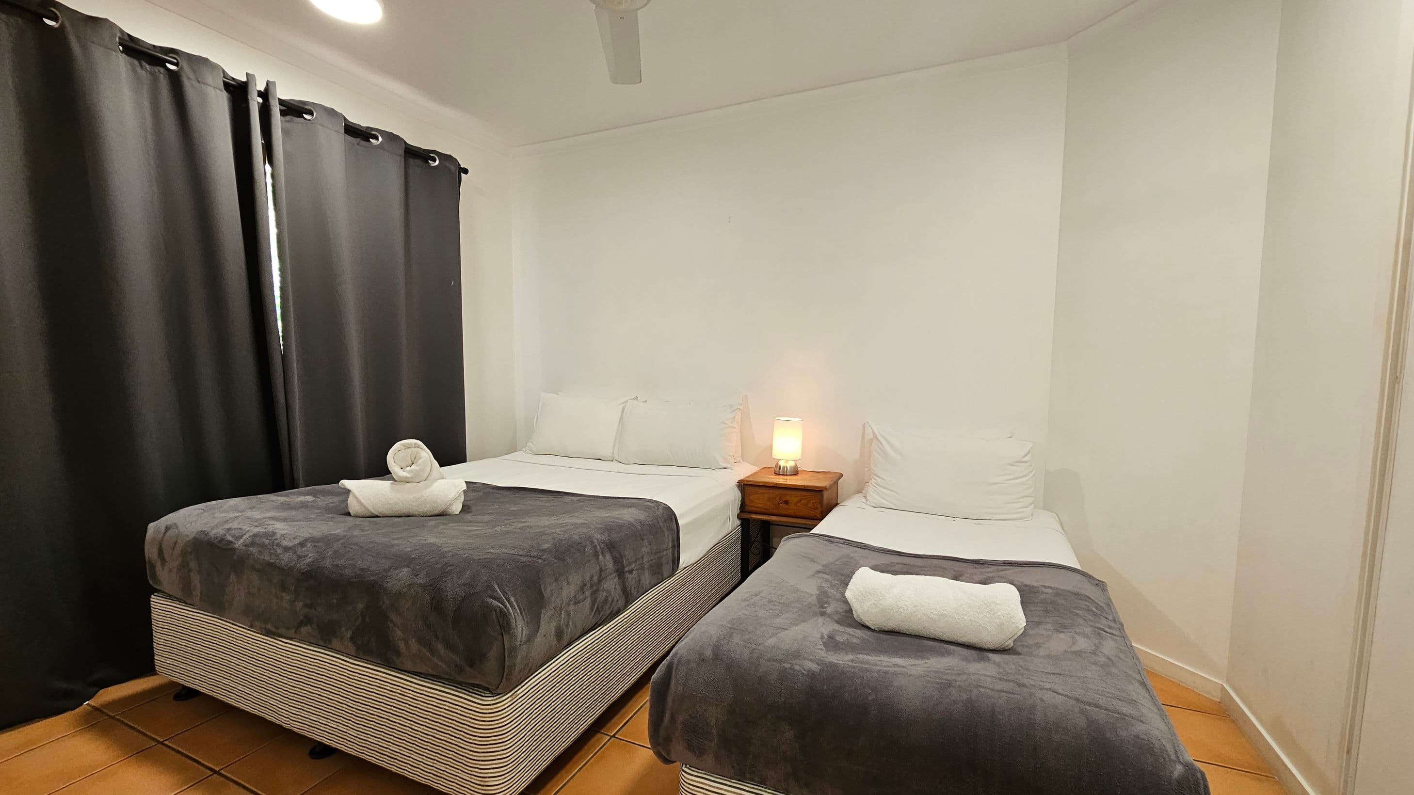 A small bedroom with two single beds, each covered with a dark blanket and a rolled white towel on top. Between the beds is a wooden nightstand with a lit lamp. The left side of the room has dark curtains covering a window. The floor is tiled with a terracotta colour, and the ceiling has a white fan.