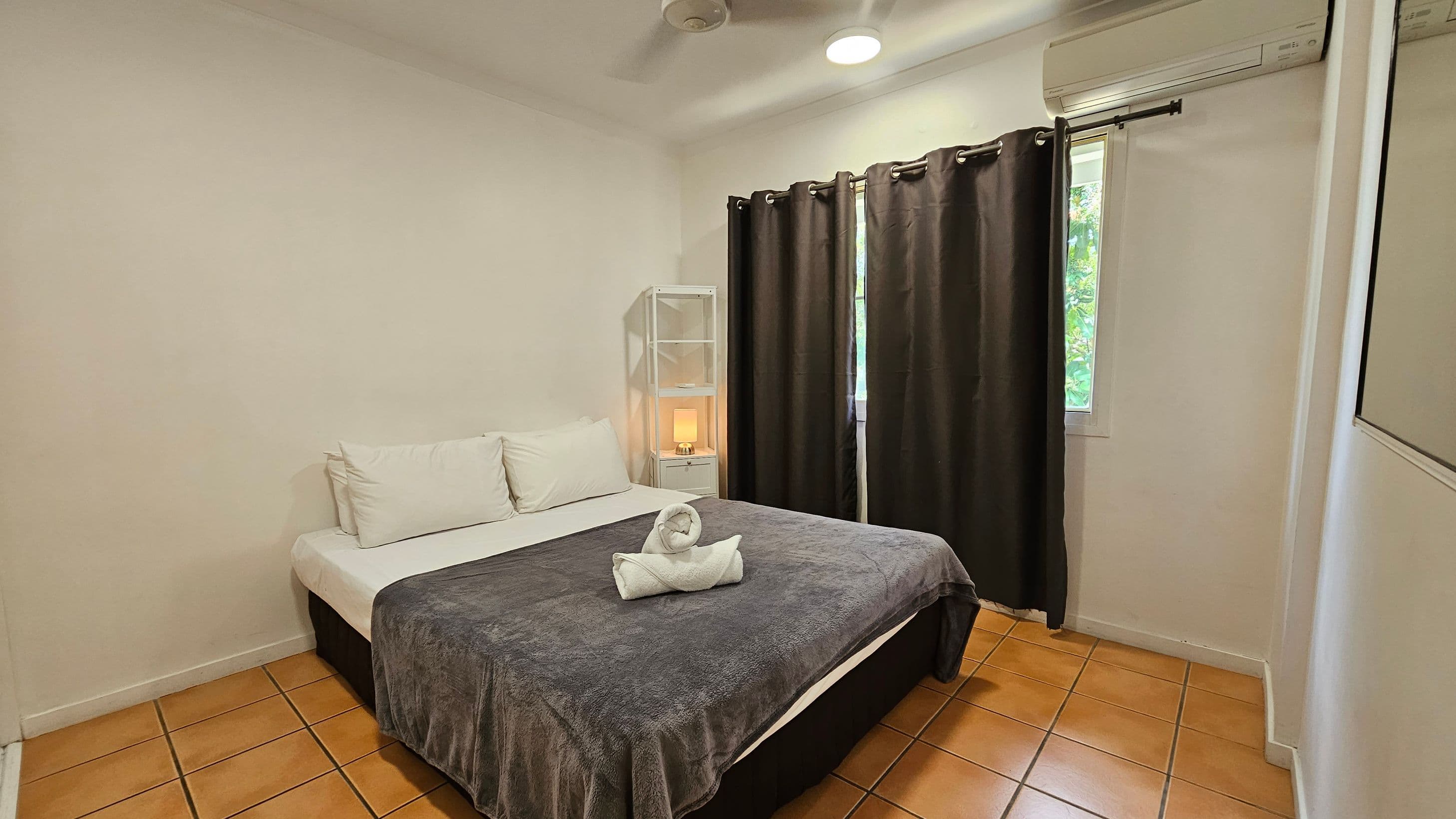 A bedroom with terracotta tiled flooring and a white ceiling. In the centre, there's a double bed with white pillows and a grey blanket, topped with a towel folded into a swan shape. To the right, a window with dark curtains and an air conditioning unit above. A white shelving unit with a lit lamp is next to the bed. A mirror is partially visible on the right wall.