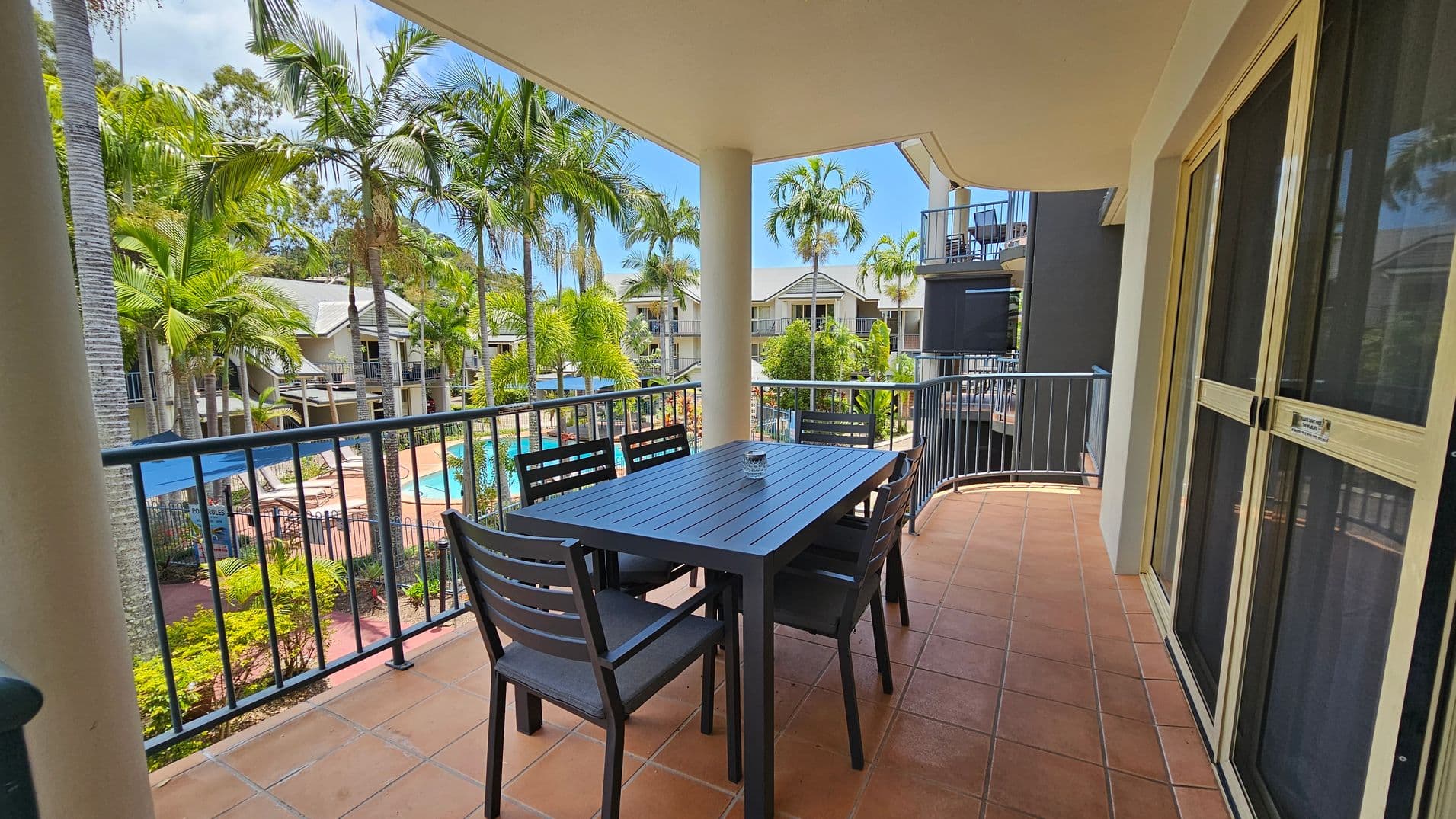 A balcony with terracotta tiles features a dark wooden table and six matching chairs. The railing overlooks a pool area surrounded by palm trees and other greenery. In the background, there are several residential buildings with balconies. On the right, sliding glass doors lead inside. The ceiling is light-coloured and flat.