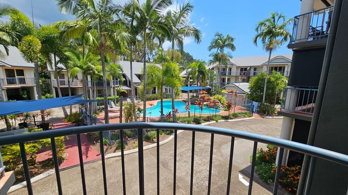 A view from a balcony overlooking a courtyard with a swimming pool surrounded by palm trees. The pool area is fenced and has sun loungers and blue shade sails. There are two-storey buildings with balconies on either side, featuring grey roofs and light-coloured walls. The ground is paved with red and grey paths, and there are landscaped gardens with various plants and flowers.