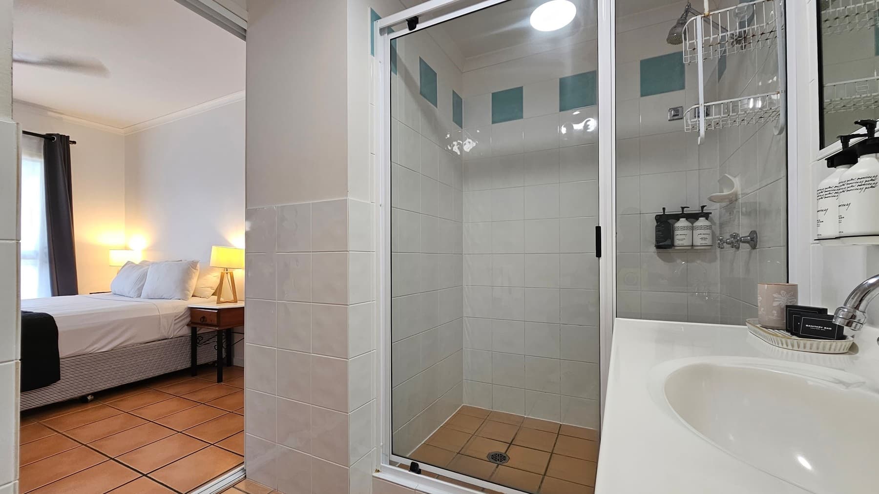 A bathroom with a glass shower enclosure in the centre, featuring white tiles and green accents. Inside the shower, there are wall-mounted toiletries. To the right, a white sink with a chrome tap and various toiletries on a tray. On the left, an open doorway leads to a bedroom with a bed, bedside table, and a lit lamp. The floor is tiled in a terracotta colour.