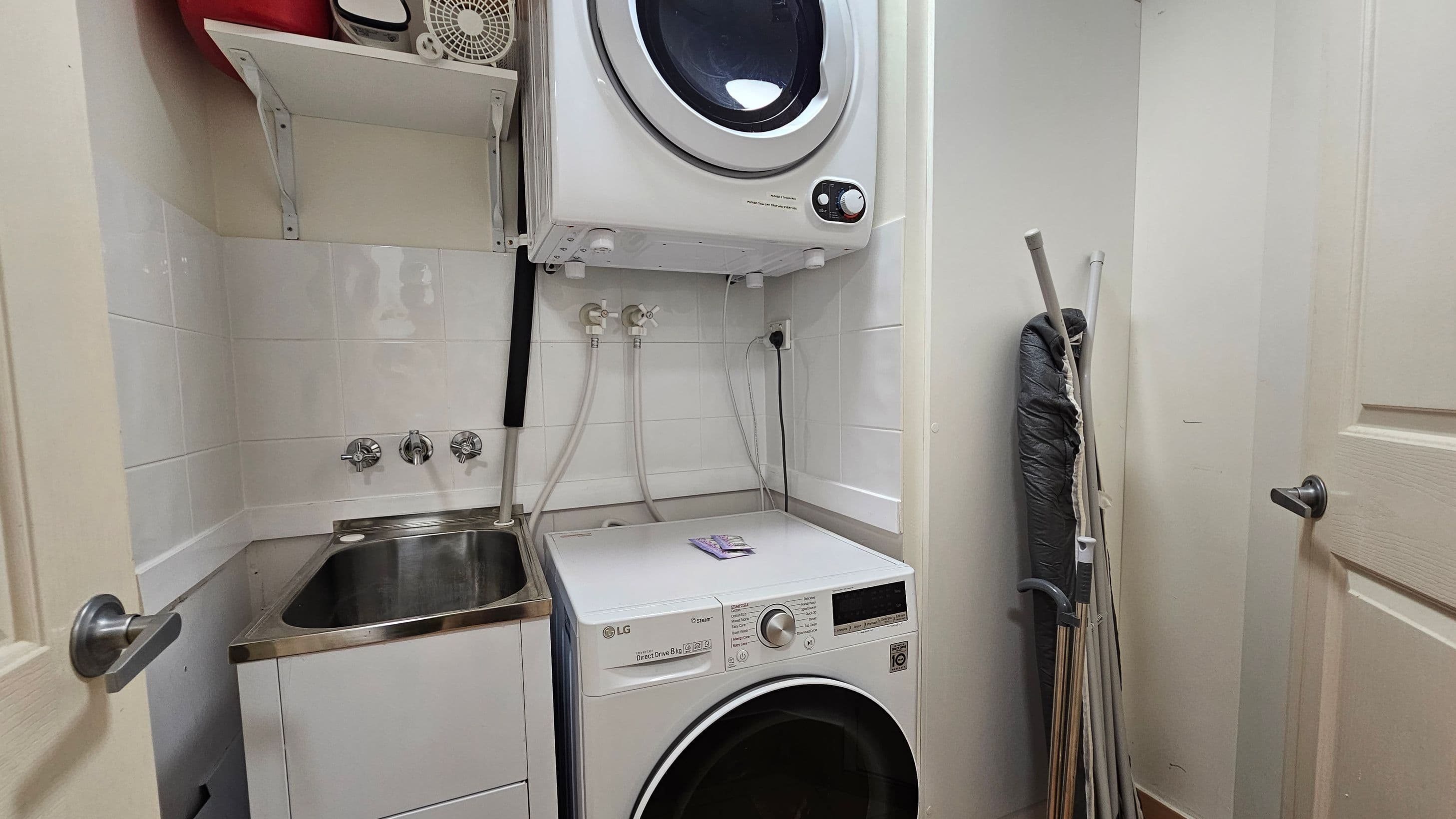 A compact laundry room with a white washing machine on the floor and a dryer stacked above it. To the left, there's a stainless steel sink with taps and a small shelf holding various items, including a fan. On the right, an ironing board and some cleaning tools are propped against the wall. The walls are tiled, and the room is enclosed by a door with a silver handle.