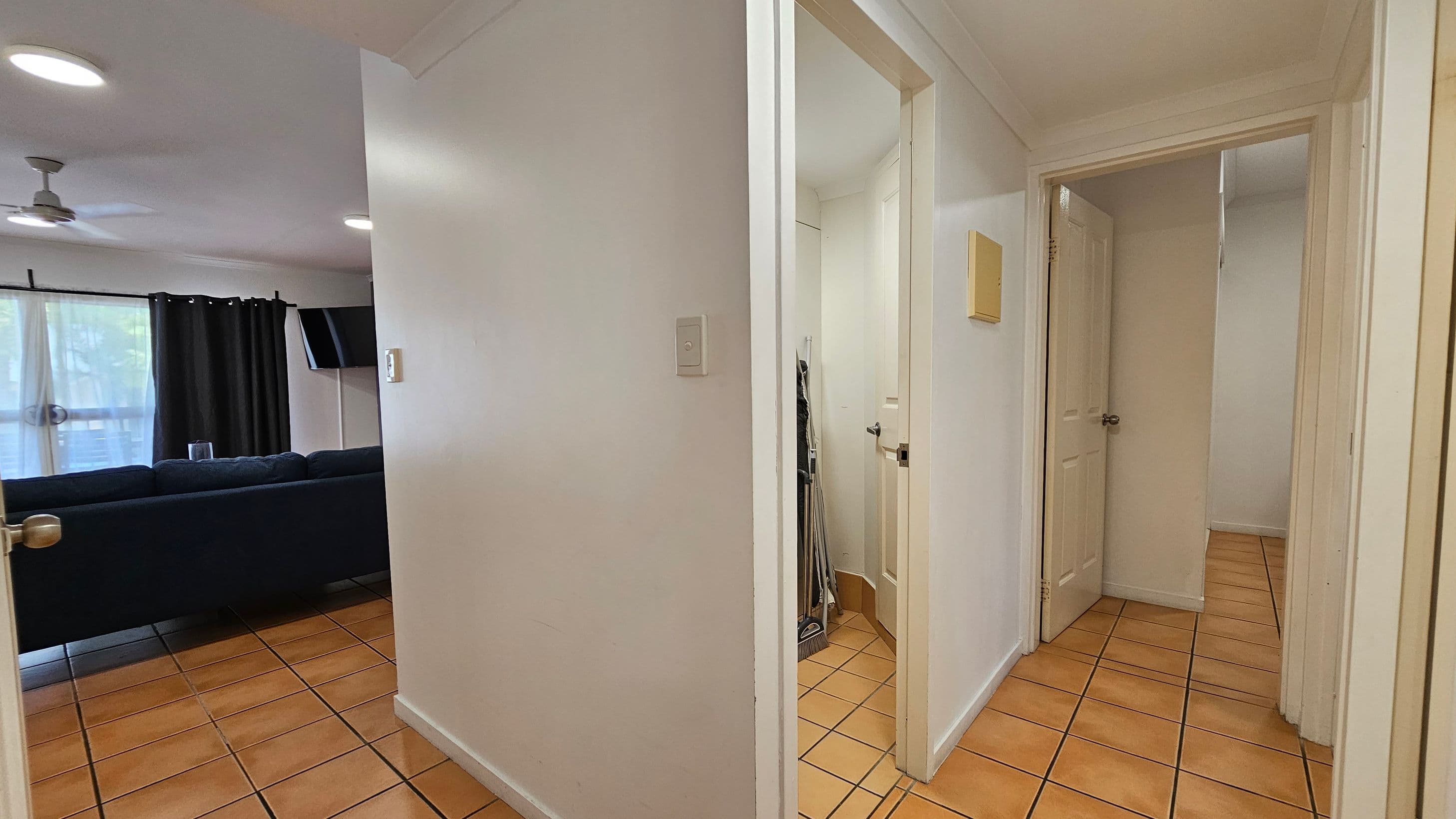 A hallway with terracotta floor tiles and white walls. On the left, an open doorway leads to a living room with a dark sofa, a wall-mounted TV, and a ceiling fan. The room has large windows with dark curtains. On the right, two open doors lead to other rooms, with cleaning tools visible in one doorway. The ceiling is white with recessed lighting.