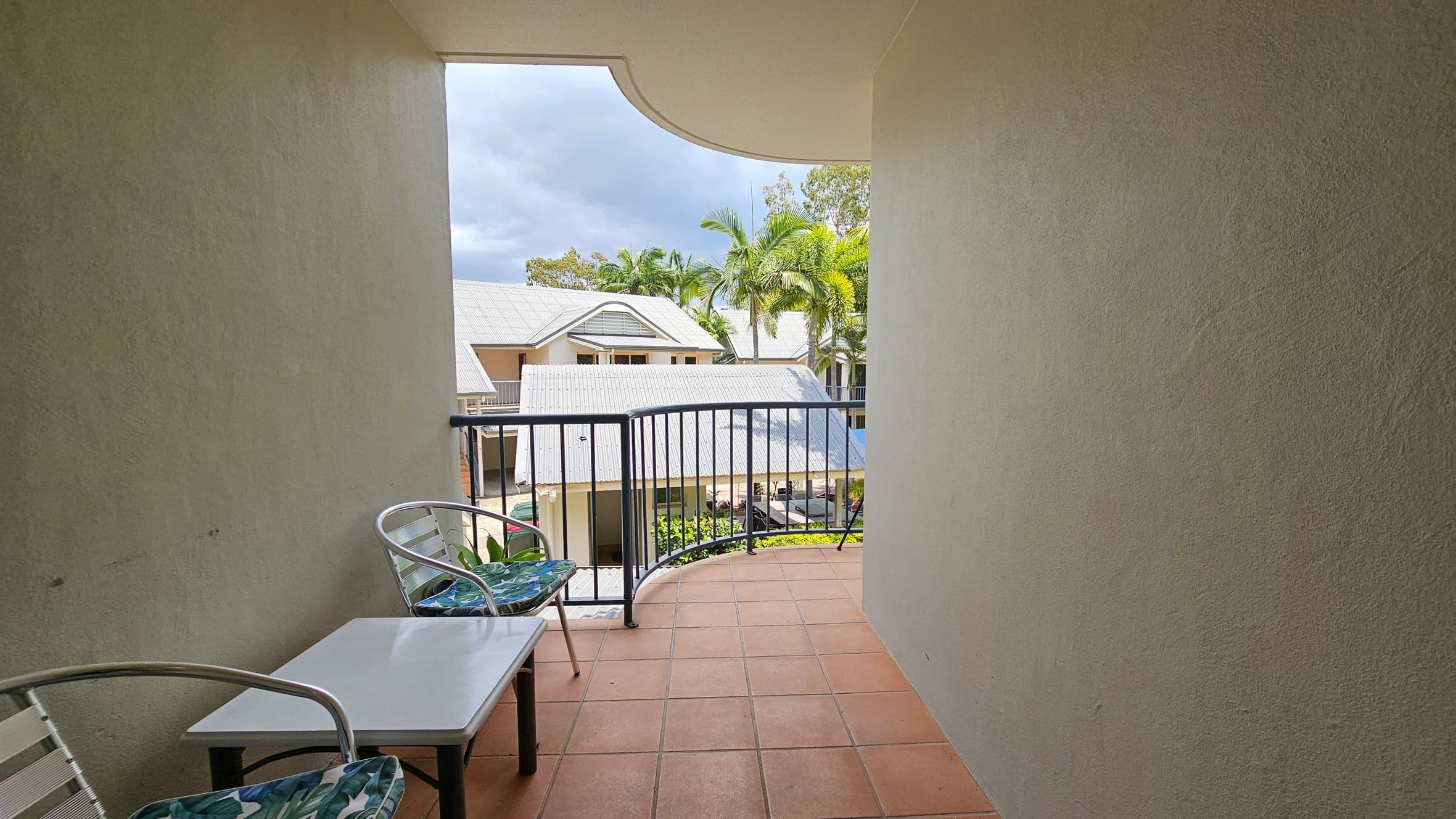 A small balcony with terracotta tiles and a black metal railing. On the left, there are two metal chairs with green patterned cushions and a small white table between them. The view overlooks a complex with light-coloured roofs and palm trees in the background. The walls of the balcony are light-coloured and textured.