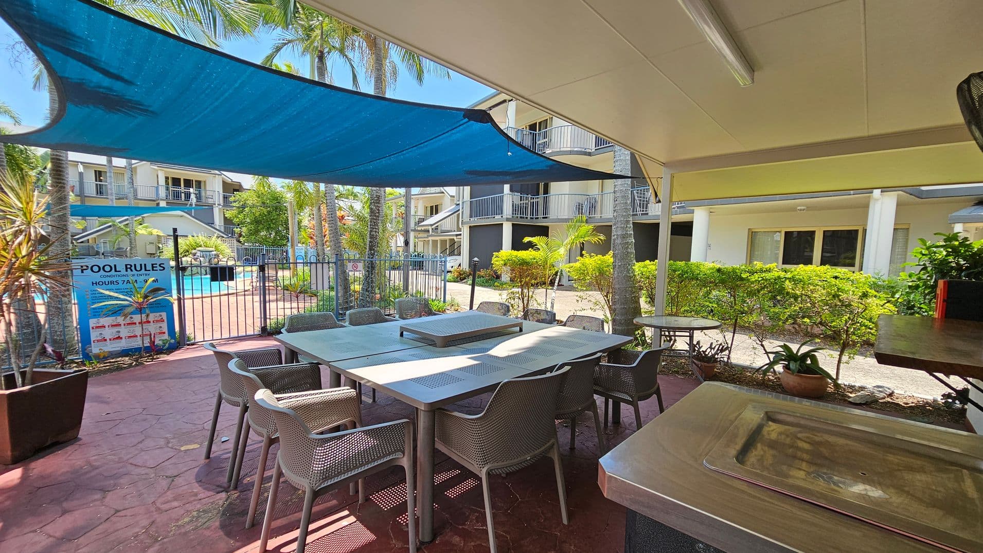 A covered outdoor area with a large metal table surrounded by chairs on a red-tiled floor. To the left, a blue shade sail provides cover, and a sign with pool rules is visible near a fenced swimming pool. In the background, there are multi-storey buildings with balconies, and palm trees and shrubs add greenery to the scene. A metal sink is in the foreground on the right.