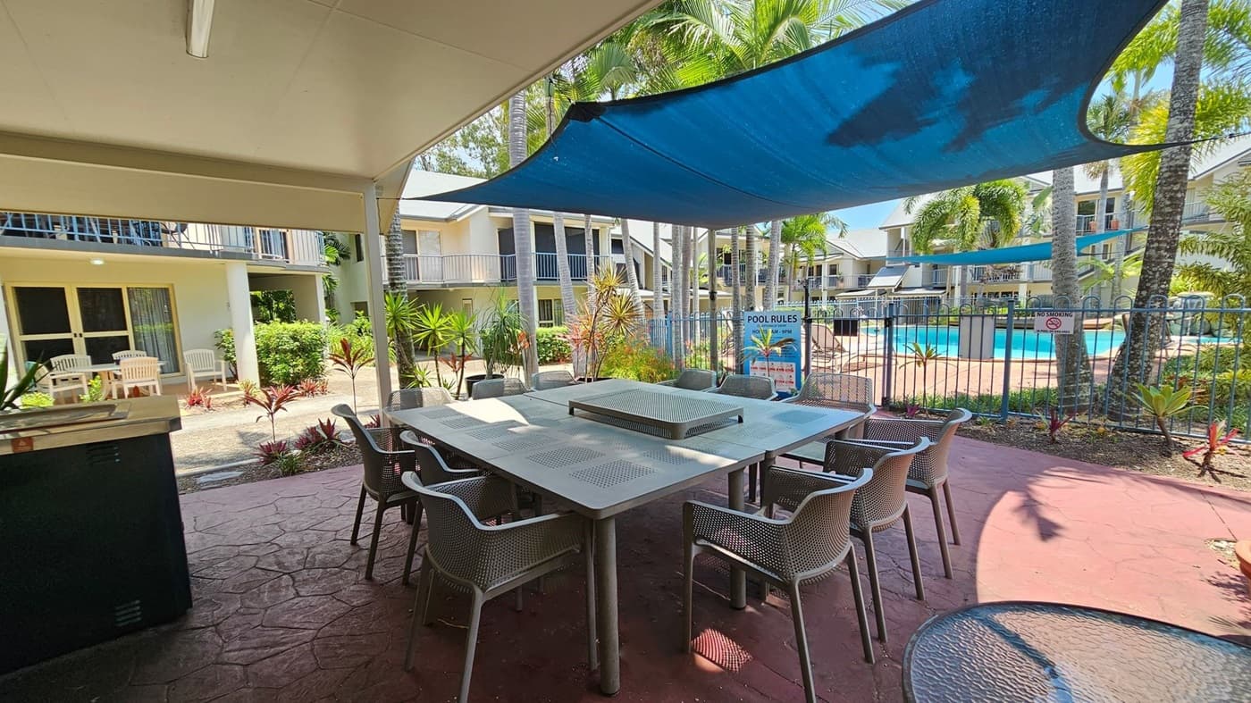 Outdoor seating area with a large square table surrounded by chairs on a red-tiled floor. A blue shade sail is overhead. In the background, there are two-storey buildings with balconies, and a fenced swimming pool area with palm trees and a sign displaying pool rules. White patio chairs are visible near the building entrances, and various plants and shrubs are scattered around the area.