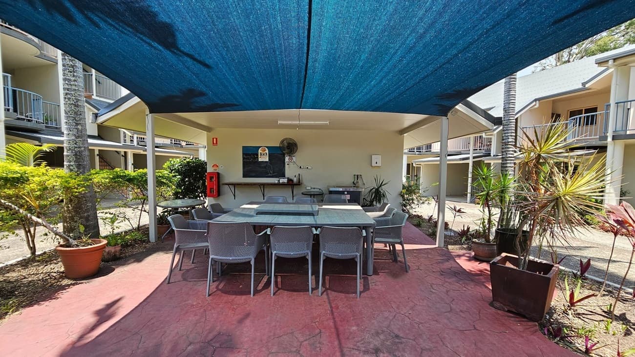 A shaded outdoor area with a large table and several chairs beneath a blue canopy. The ground is paved with red stone. In the background, there's a wall with a chalkboard, a fan, and a fire extinguisher. Potted plants and small trees are on either side. Surrounding the area are two-storey buildings with balconies.