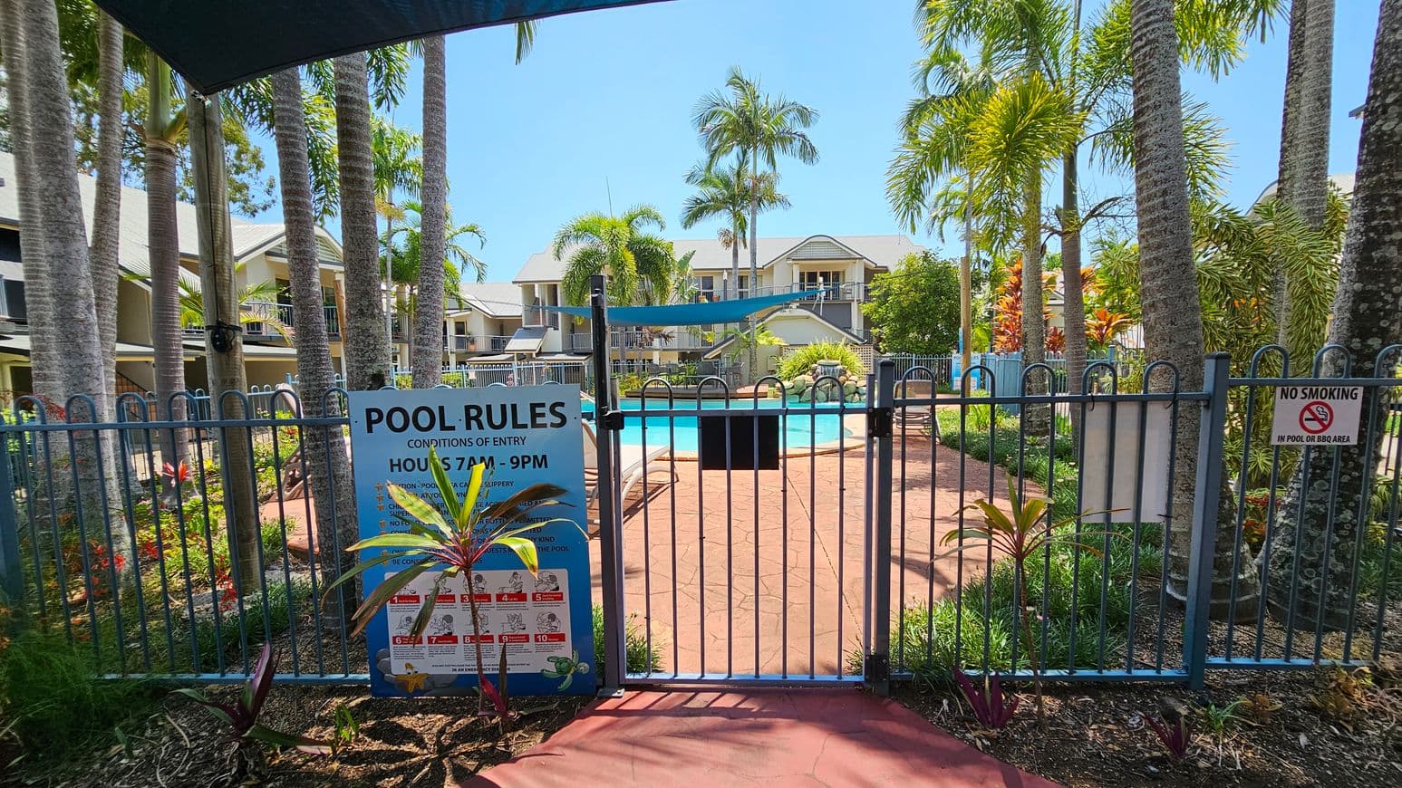 A fenced pool area with a blue gate in the foreground, displaying a "Pool Rules" sign and a "No Smoking" notice. The path leading to the pool is surrounded by lush greenery and tall palm trees. In the background, there are residential buildings with balconies overlooking the pool. The pool is visible, with sun loungers around it, and a shade sail above. The ground is paved with red tiles.