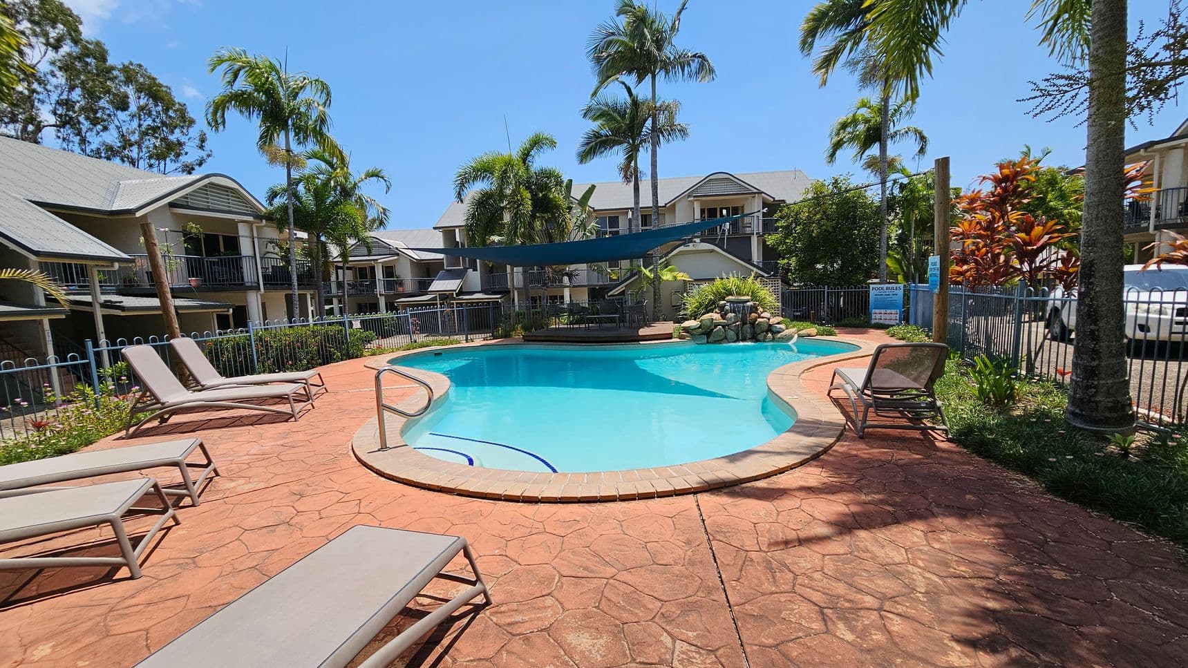 A swimming pool with a curved shape is surrounded by a red-tiled patio. Several sun loungers are placed around the pool. In the background, there are two-storey residential buildings with grey roofs and balconies. Tall palm trees and other tropical plants are scattered around the area. A blue shade sail is stretched over part of the pool, and a small rock waterfall feature is visible at the far end. A white vehicle is parked to the right, near a fence.