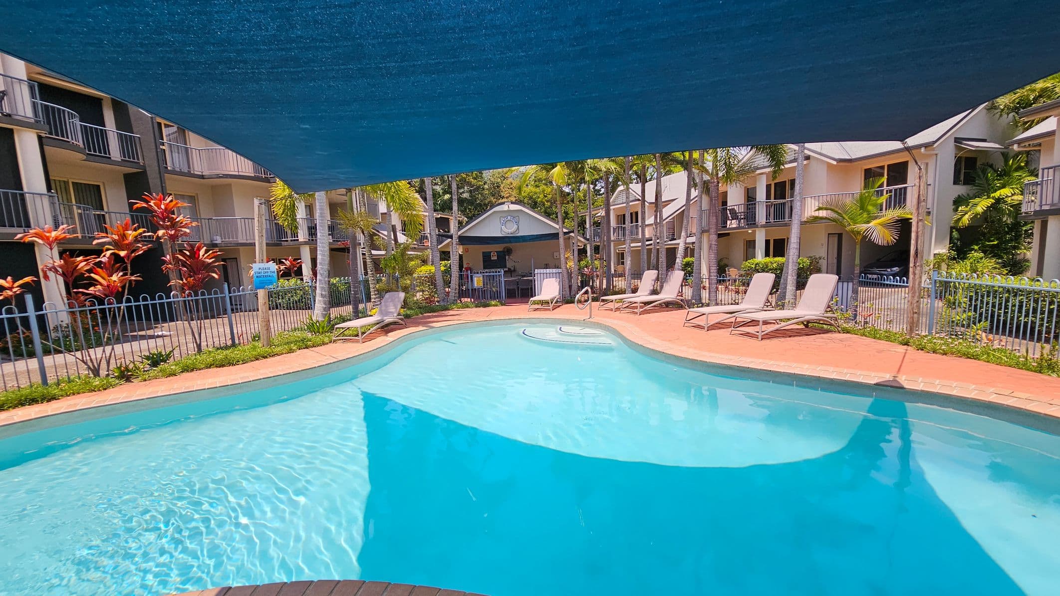 A swimming pool with clear blue water is in the foreground, surrounded by a red brick deck. Several sun loungers are placed along the right side. In the background, there are two-storey buildings with balconies, and a small structure with a clock. Tall palm trees and red-leaved plants are scattered around the area. A blue shade cloth is stretched above the pool, providing partial cover. A metal fence encloses the pool area.