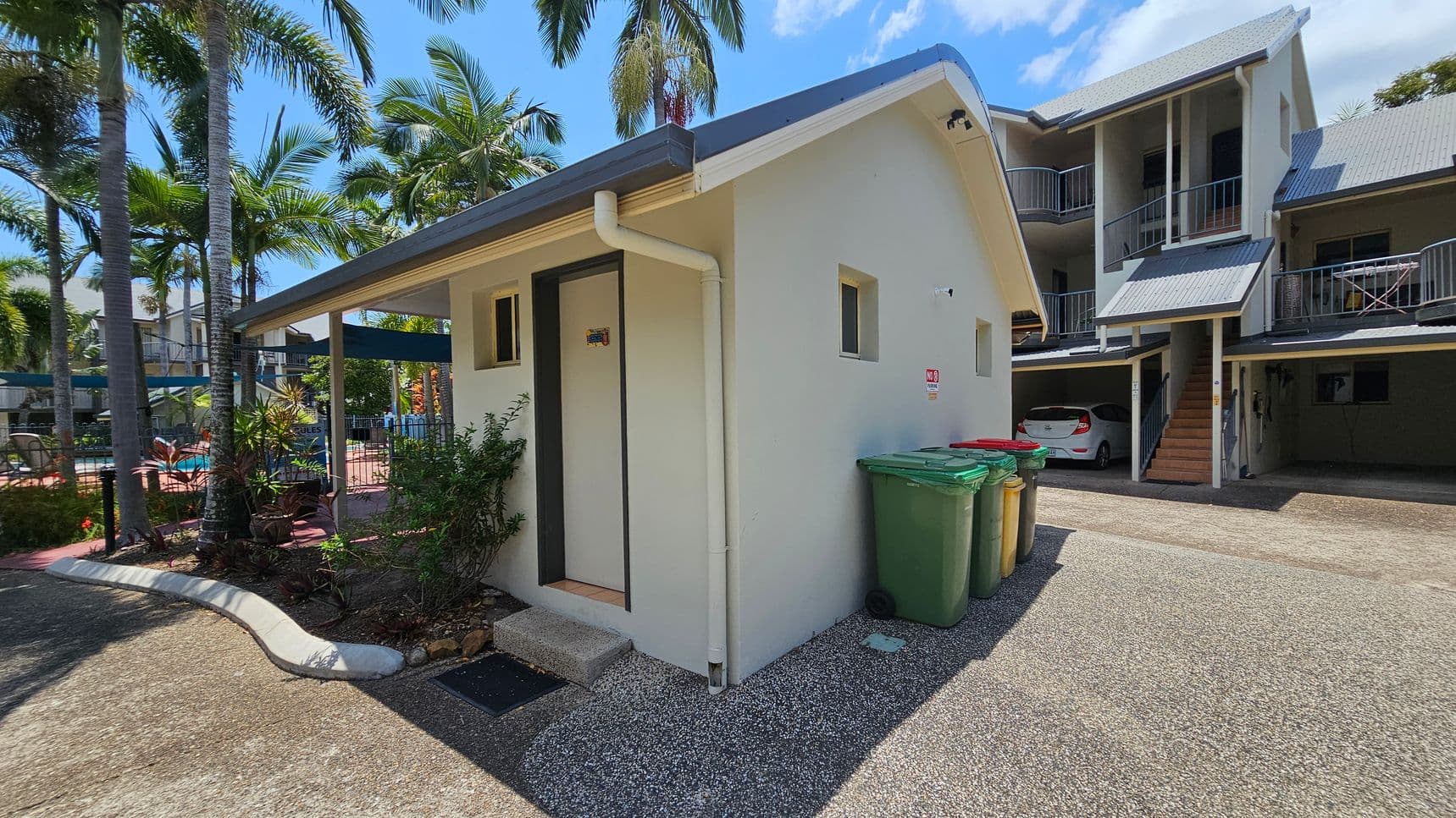 A small, light-coloured building with a sloped roof is in the foreground, featuring a door and two small windows. Green and yellow bins are placed against its side. To the right, a larger building with a grey roof and balconies is visible, with a white car parked underneath. On the left, there are palm trees and a glimpse of a swimming pool area with a fence and sun loungers. The ground is a mix of concrete and gravel.