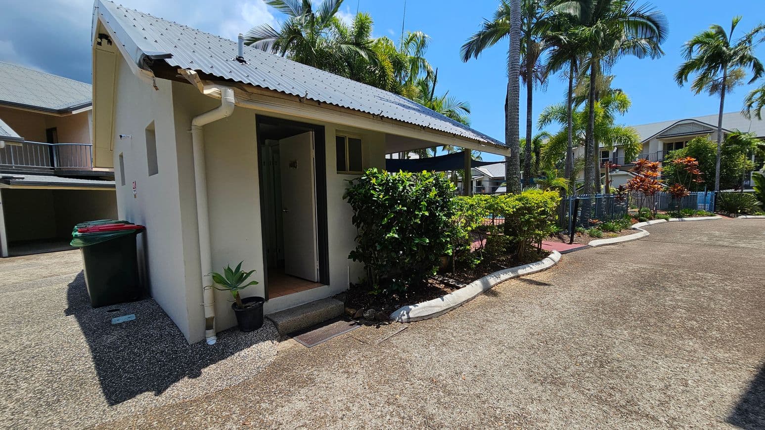 A small building with a corrugated metal roof is on the left, featuring a partially open door and two small windows. A black bin with a red lid and a green bin are beside the building. A potted plant sits near the entrance. On the right, a pathway lined with lush green bushes and tall palm trees leads to a residential area with houses in the background. The ground is covered with gravel, and the area is well-lit by sunlight.