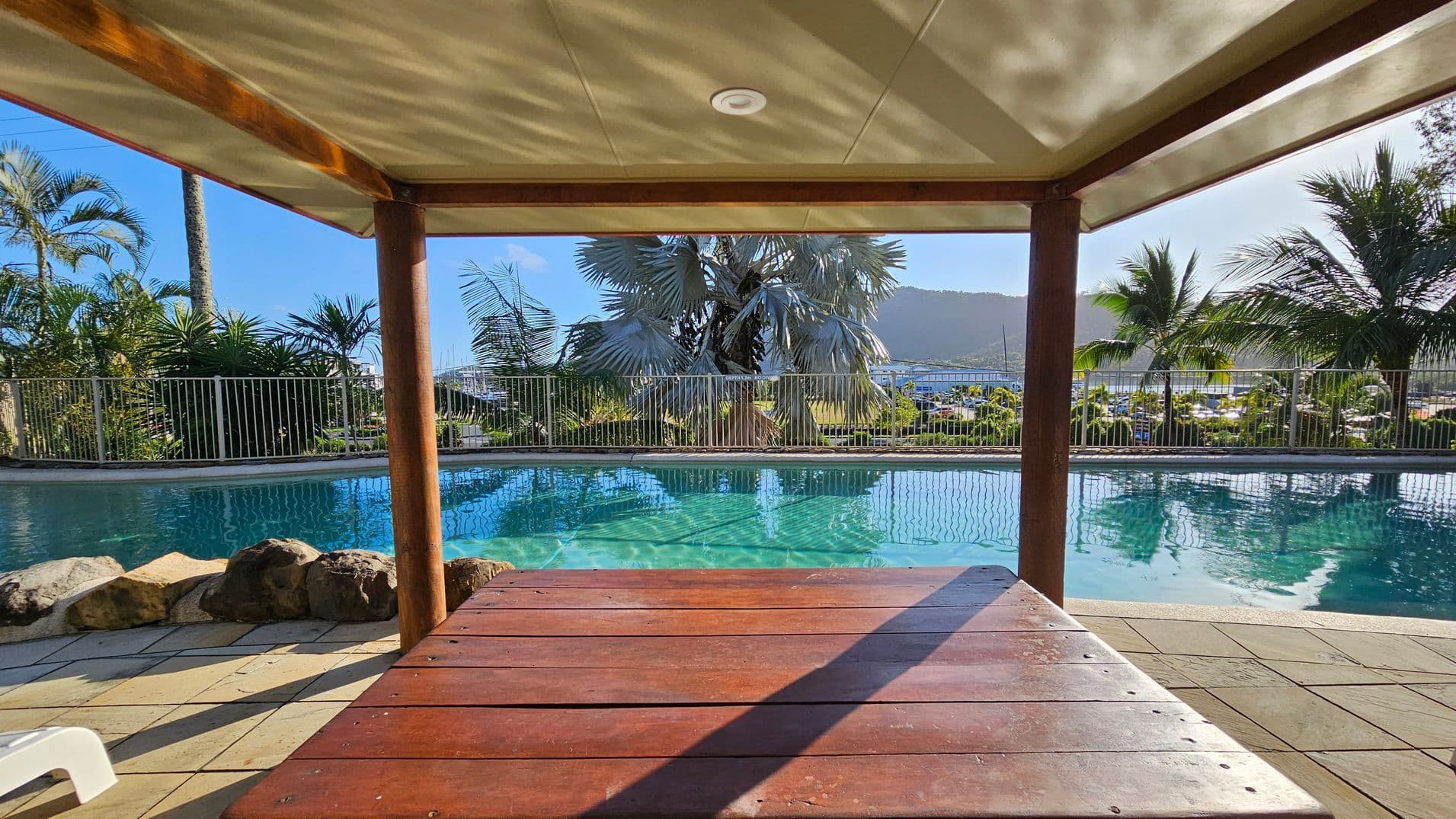 A gated pool area with a corrugated metal roofed shelter in the centre. The gate has signs displaying pool rules and a notice about no glass allowed. Lush greenery, including palm trees and various plants, surrounds the area. Beyond the gate, a glimpse of the pool is visible. The ground is paved with small stones, and the sun is shining brightly, casting shadows.