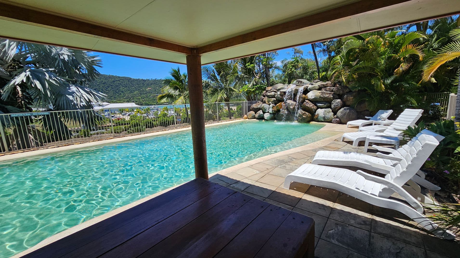 A swimming pool with clear water is surrounded by a tiled deck. On the right, several white sun loungers are lined up. In the background, a rock formation with a waterfall is visible, along with lush green palm trees and foliage. A metal fence runs along the left side, with a view of a marina and hills in the distance. The foreground shows a shaded area with a wooden table.