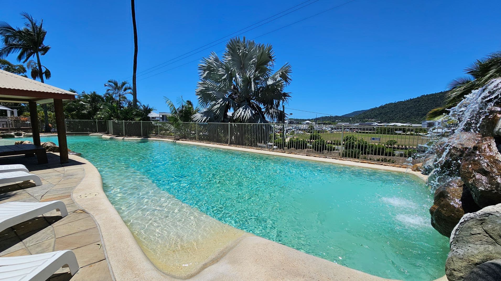 A swimming pool with clear blue water is in the centre, featuring a small waterfall on the right cascading over rocks. On the left, there are several white sun loungers on a tiled patio under a shaded area. In the background, there are palm trees and a metal fence, with a view of a distant hillside and some buildings. The sky is clear and blue.