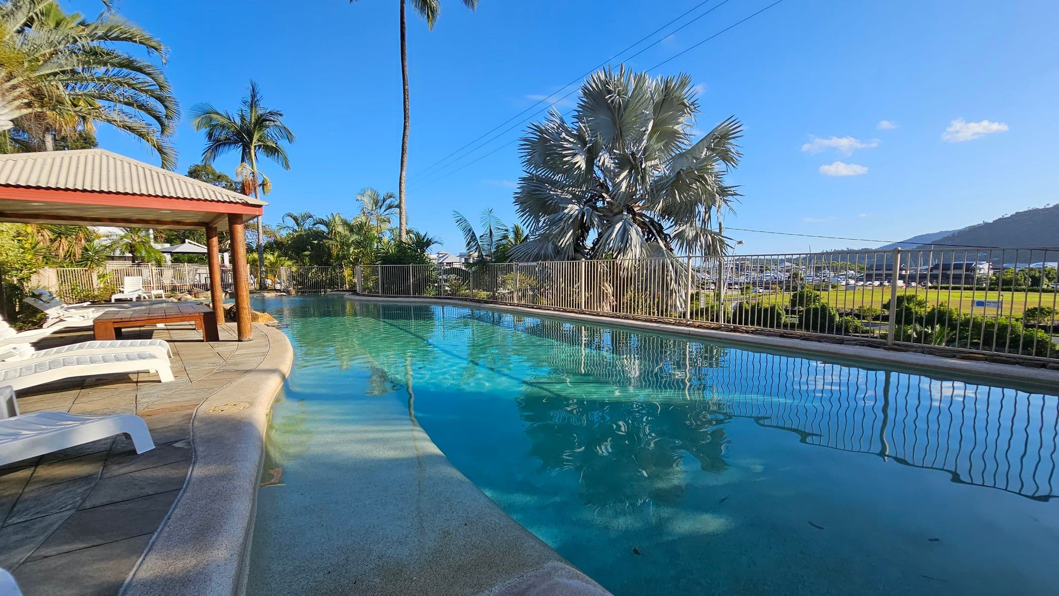 A swimming pool with clear blue water is on the right, bordered by a metal fence. On the left, there is a shaded area with a wooden structure and several white sun loungers on a tiled floor. Tall palm trees and lush greenery surround the pool area. In the background, there is a view of a grassy field and a marina with boats, set against a backdrop of hills.