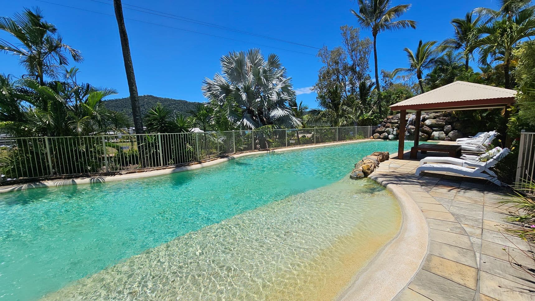 A swimming pool with clear turquoise water is surrounded by a stone-tiled deck. On the right, several white sun loungers are positioned under a wooden gazebo with a corrugated roof. In the background, lush tropical plants and palm trees line the perimeter, with a metal fence separating the pool area from the greenery. A hill covered in vegetation is visible in the distance.