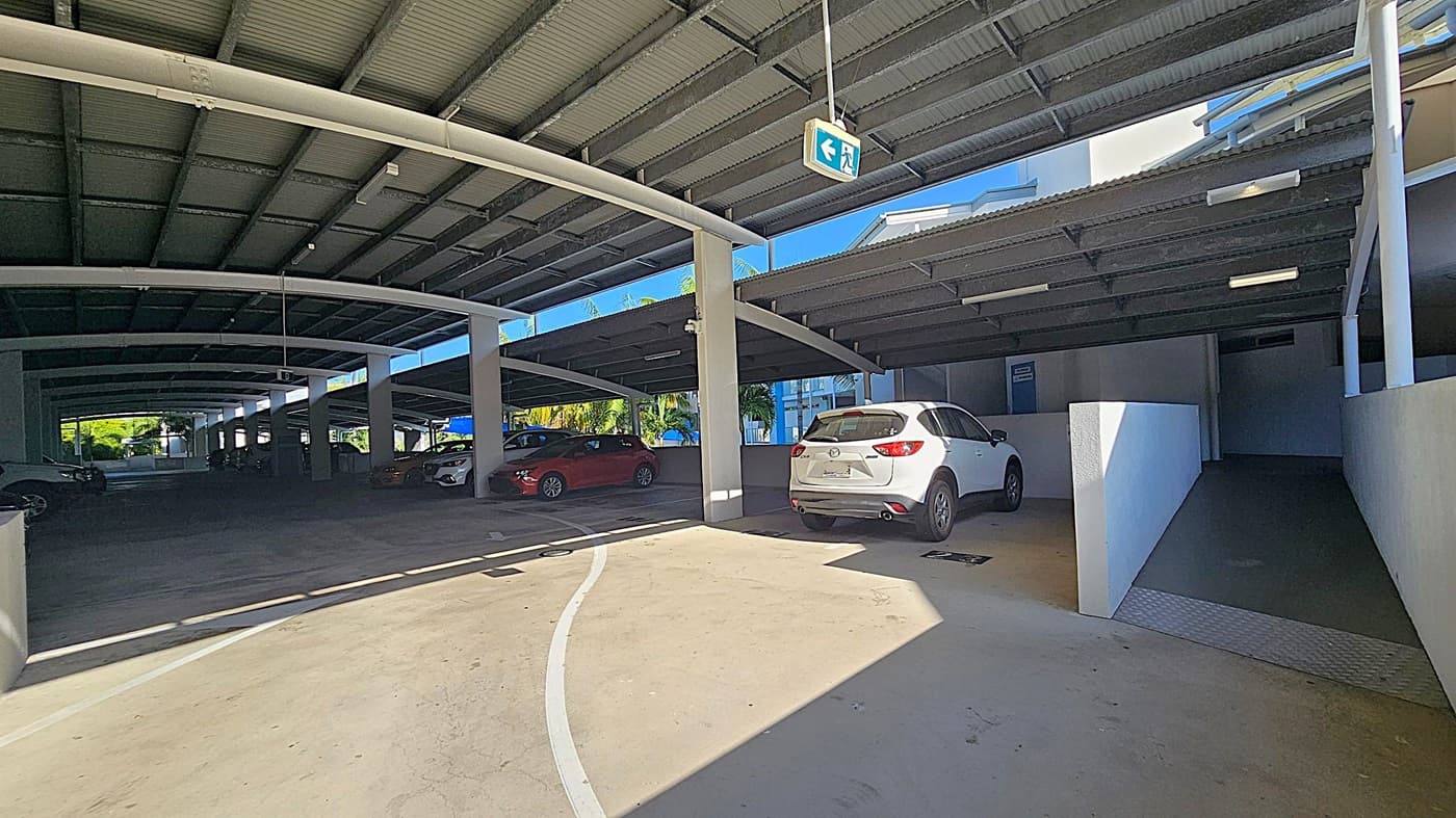 A concrete driveway curves upwards on the left, bordered by a tall, dense hedge. On the right, a covered parking area is visible, with a sign listing car parking units. The ceiling of the parking area is textured, and the floor is smooth concrete. Sunlight casts shadows across the driveway and parking area.