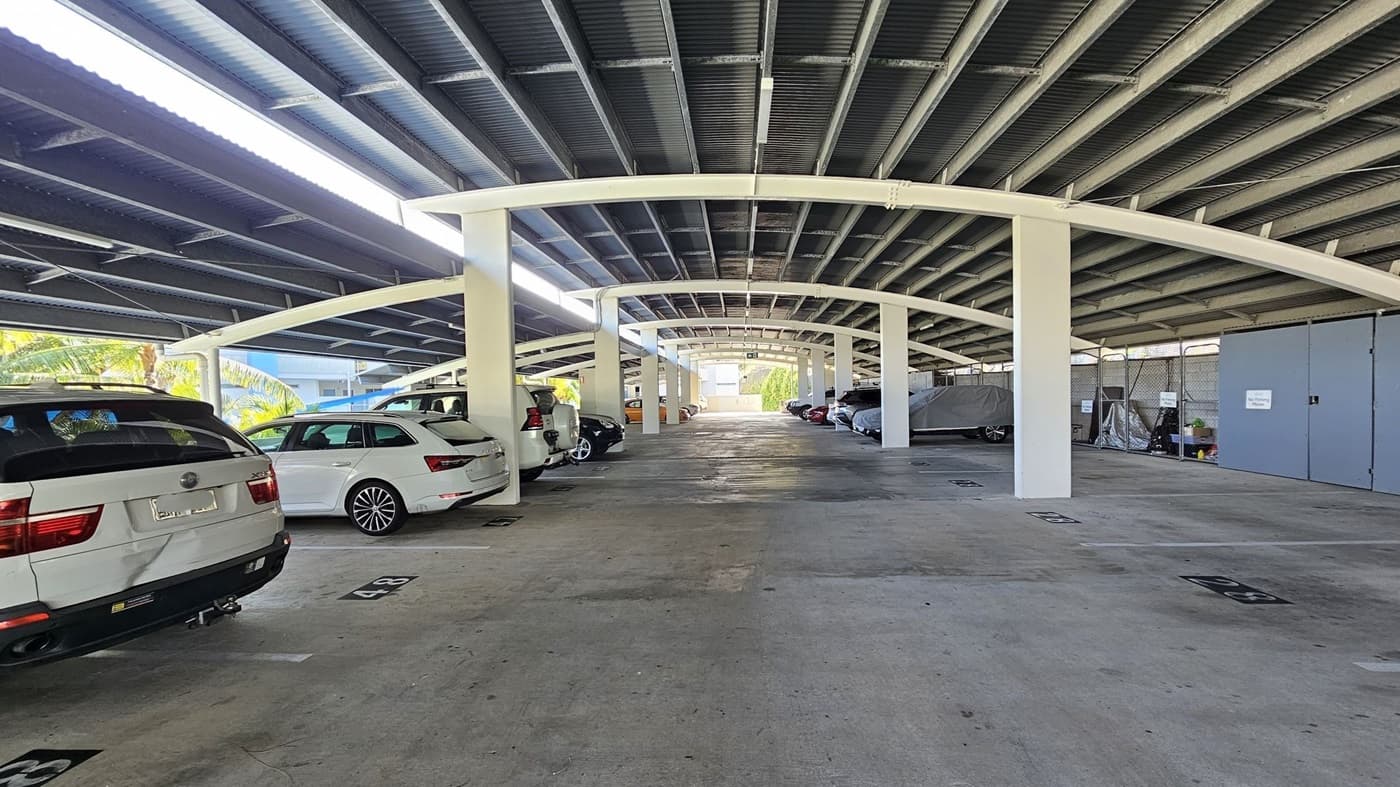 A covered car park with a concrete floor and a metal ceiling. On the left, several parked cars, including a white SUV and a white estate car. On the right, more parked cars, some covered, and a blue door with signs. The area is supported by white pillars, and there are parking space numbers marked on the ground. In the background, there is an open area with natural light coming in.