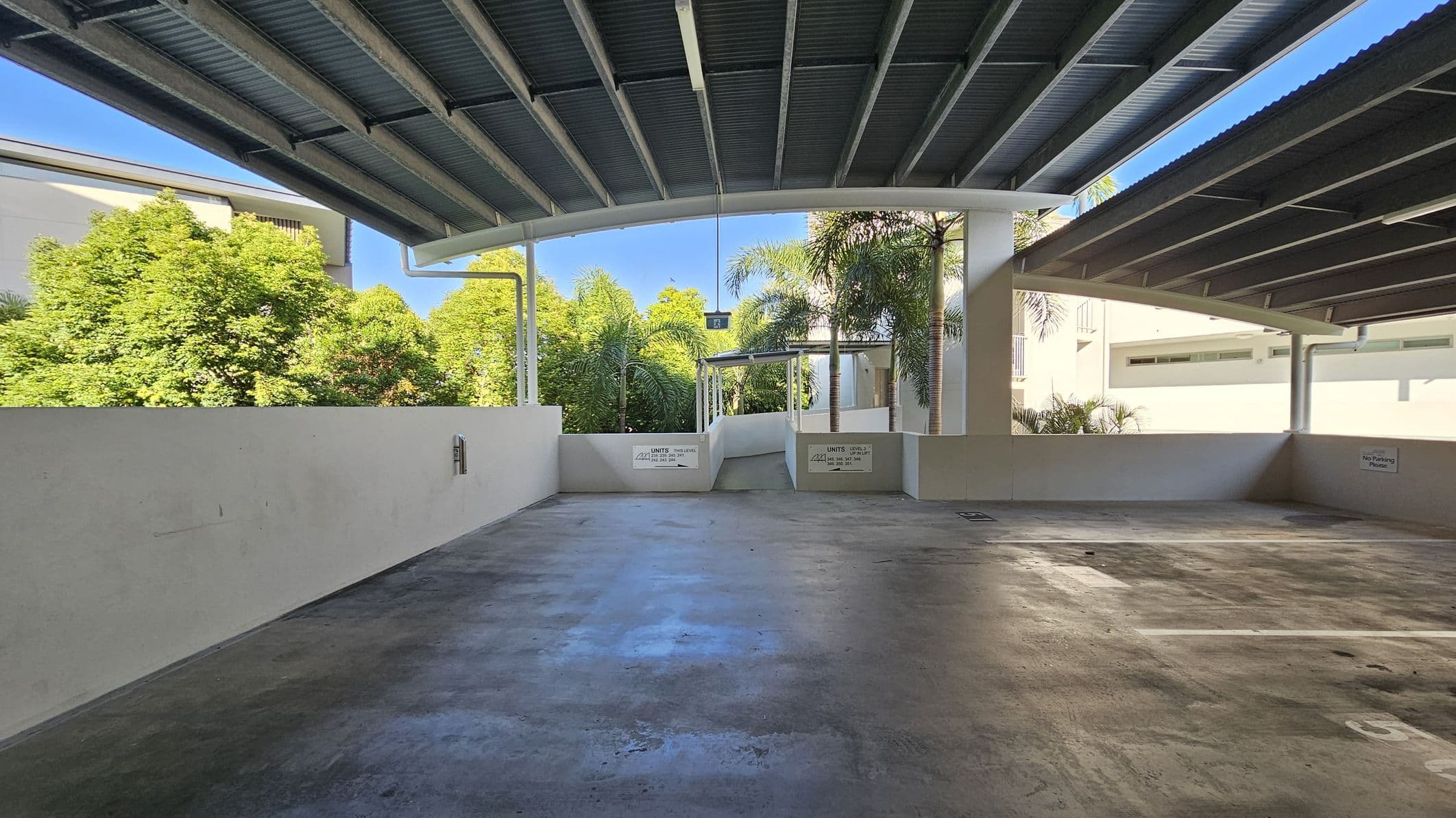 A covered parking area with a concrete floor and a corrugated metal ceiling. In the background, there are lush green trees and a building partially visible on the left. The parking area is open on the sides, allowing natural light to illuminate the space. Signs on the walls indicate reserved parking for specific units. The area is clean and well-maintained, with clear parking lines on the ground.