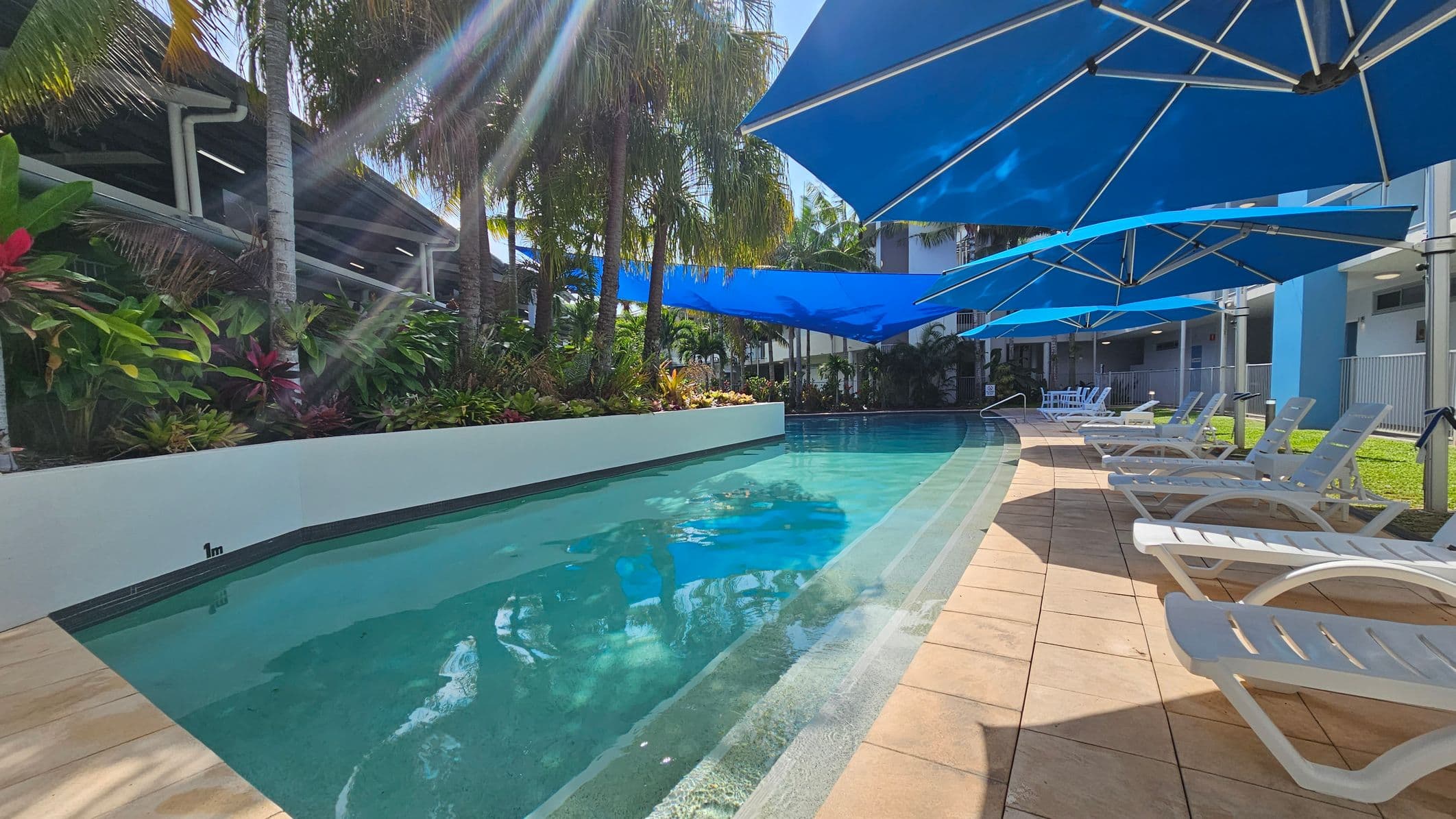 A swimming pool with clear water is surrounded by a tiled deck. On the right, there are several white sun loungers under large blue umbrellas. To the left, lush green plants and palm trees line the pool's edge. A sign with pool rules is visible on the left wall. The area is well-lit, suggesting a sunny day.