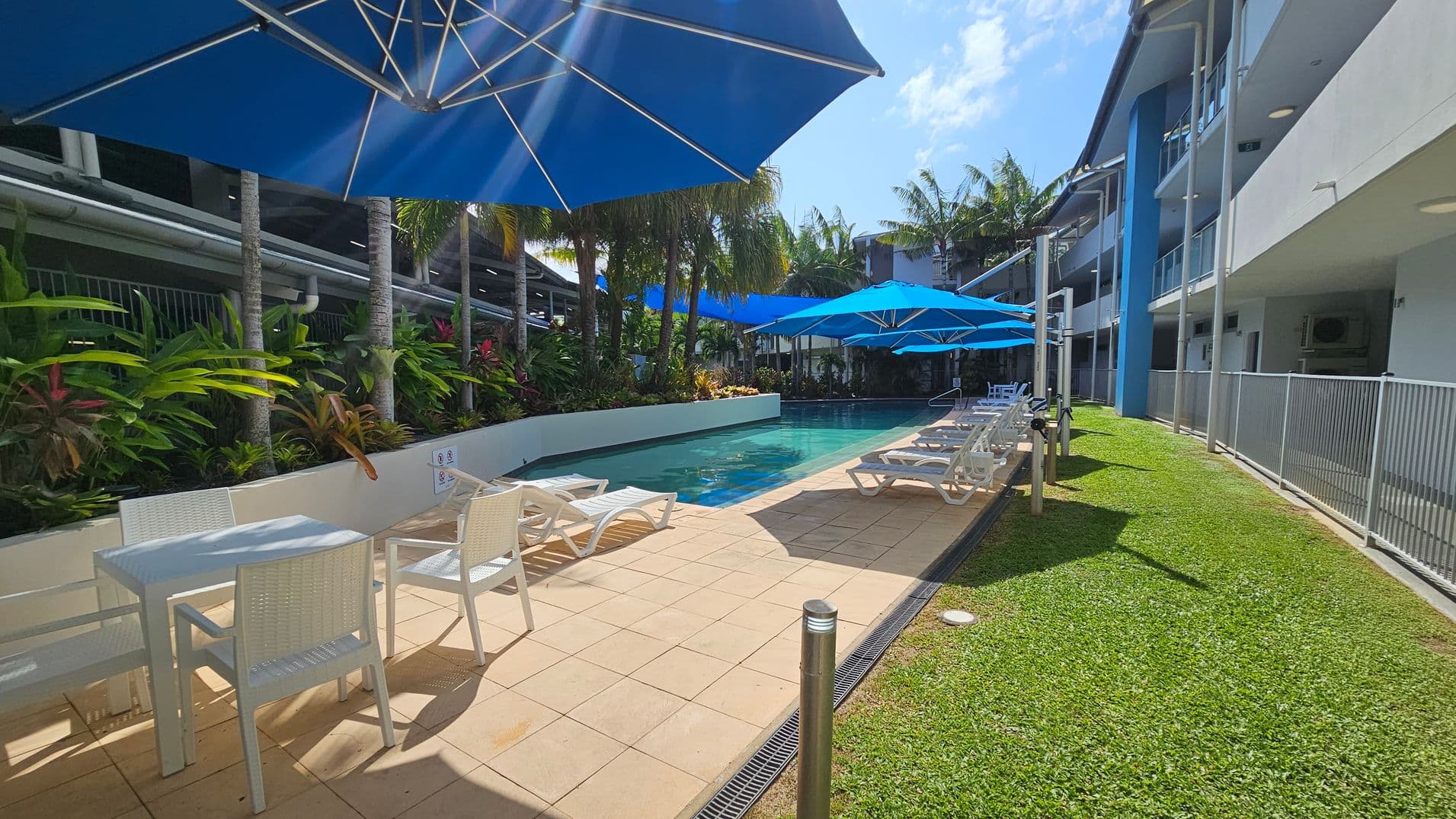 A swimming pool area with blue umbrellas providing shade over white tables and chairs on the left. Sun loungers are lined up along the poolside. The pool is surrounded by lush green plants and palm trees. On the right, there's a grassy area and a building with balconies and a blue accent wall. The floor is tiled, and a metal railing separates the grass from the walkway.