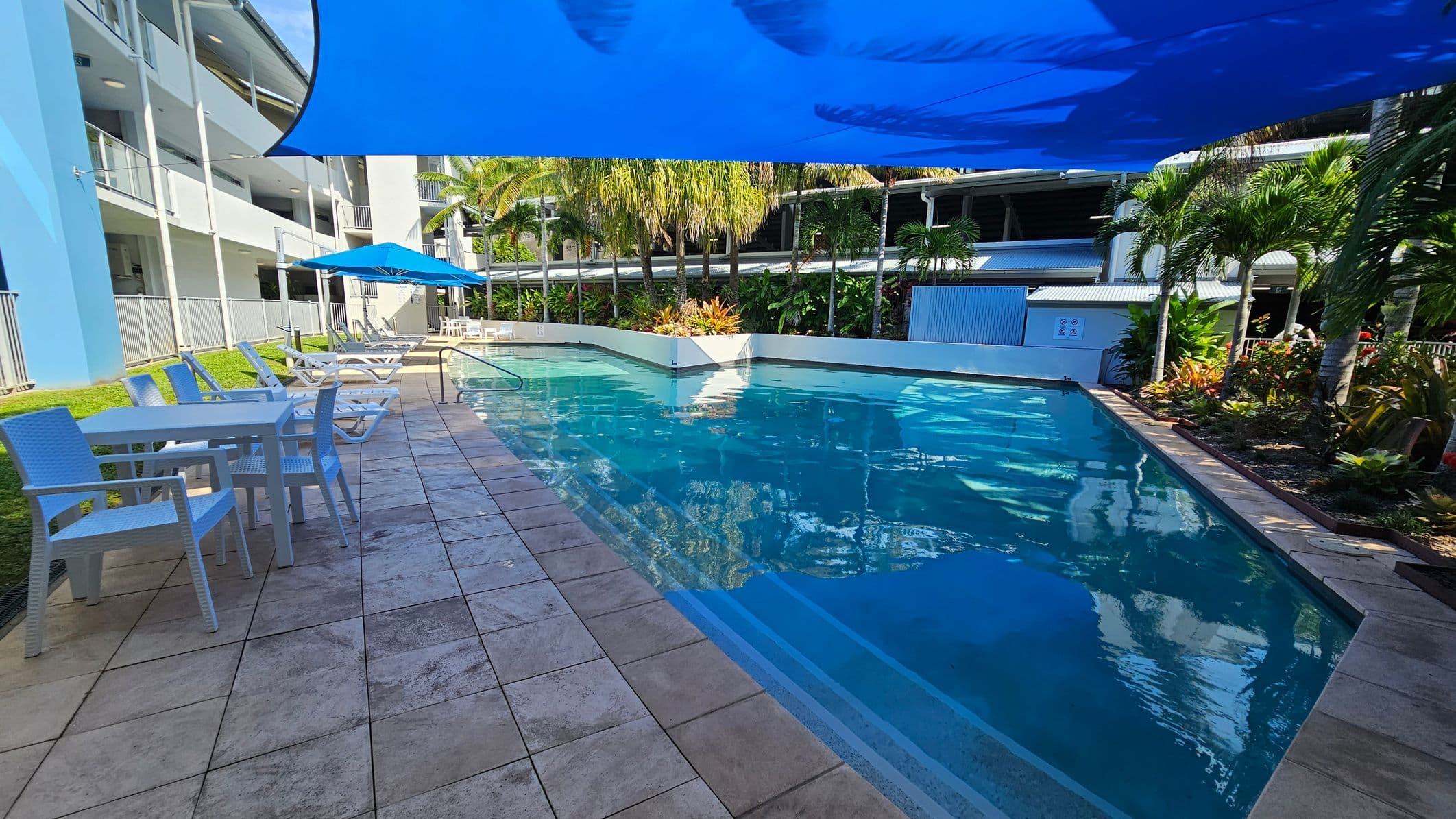 A swimming pool with clear blue water is surrounded by a tiled deck. On the left, there are white tables and chairs, and several sun loungers under a blue umbrella. A blue shade sail covers part of the area. In the background, there are tall palm trees and lush greenery. A multi-storey building with balconies is visible on the left side.
