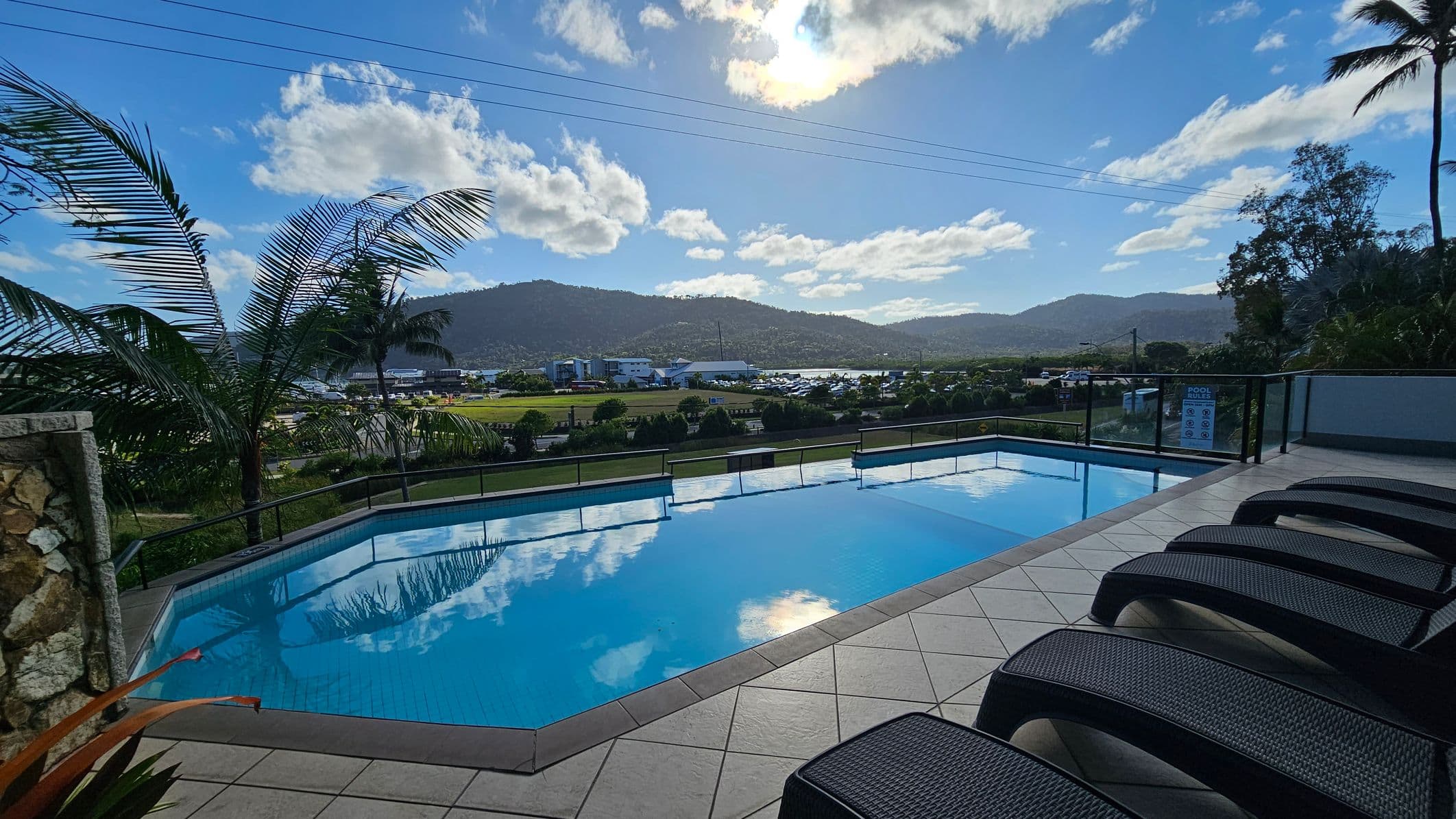 A swimming pool with clear blue water is in the foreground, surrounded by a tiled deck. On the right, several dark-coloured sun loungers are lined up. To the left, palm trees sway gently. In the background, a landscape of green hills and a small town is visible, with a few scattered buildings and parked vehicles. The scene is bordered by a glass railing, and the sun is partially obscured by clouds, casting reflections on the pool's surface.