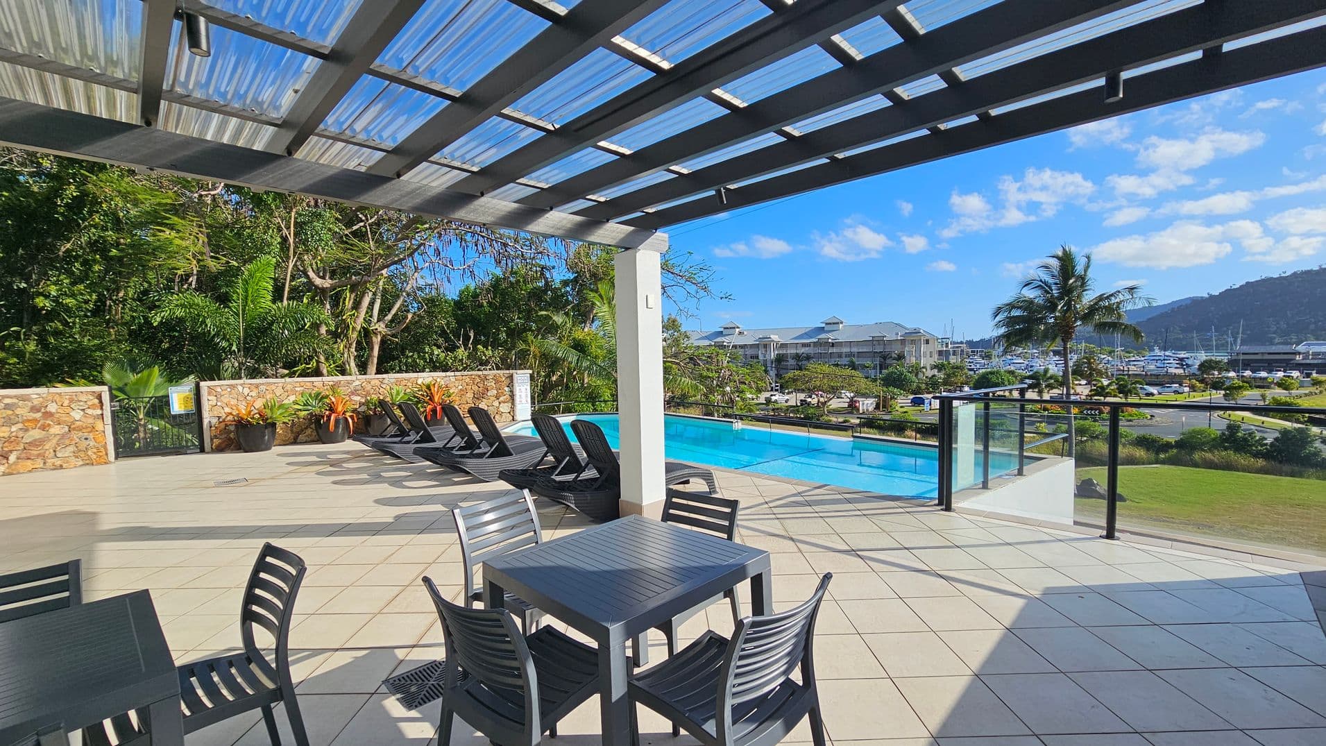 A patio area with a tiled floor and a transparent roof. On the left, there is a table with four black chairs. In the middle, several black sun loungers are arranged near a stone wall with potted plants. On the right, a swimming pool is visible, bordered by glass railings. Beyond the pool, there is a view of a marina with boats and a building in the background, along with a palm tree and grassy area.