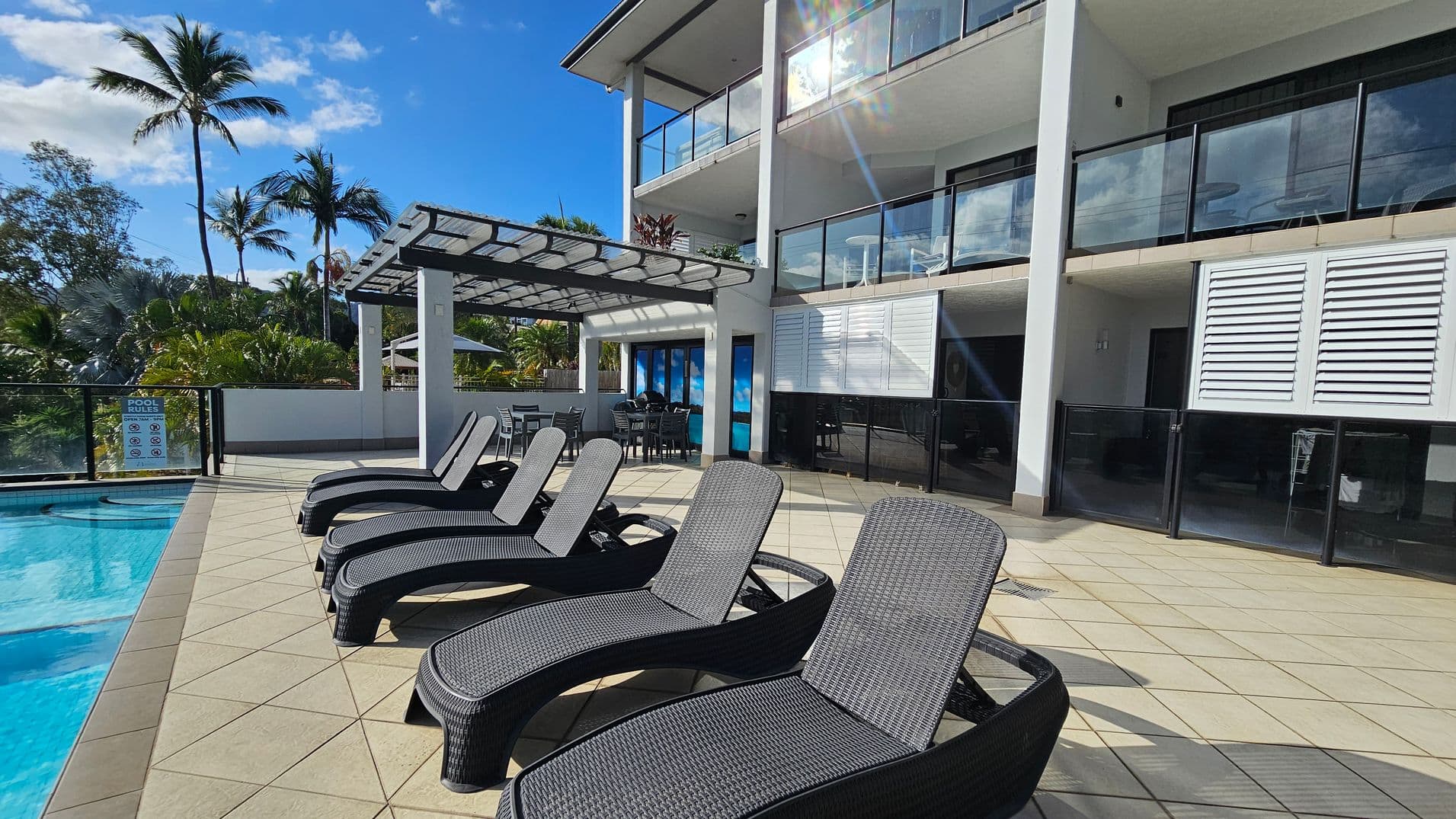 Poolside area with several black wicker sun loungers on a tiled patio. To the left, a clear blue swimming pool is visible. In the background, a modern building with glass balconies and white walls stands tall. A pergola with a dining table and chairs is situated near the building. Tall palm trees and lush greenery surround the area, adding a tropical feel. The sky is clear and blue.