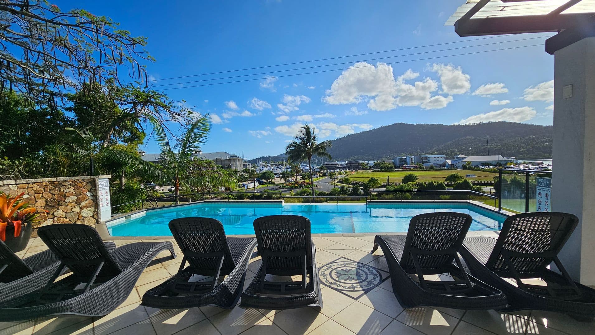 Five black sun loungers are arranged on a tiled patio in the foreground, facing an infinity pool. To the left, a stone wall with tropical plants and trees. In the background, a scenic view of a marina with boats, a grassy area, and a mountain range. A palm tree stands near the pool's edge, and a building is partially visible on the right. The pool area is enclosed by a glass railing.