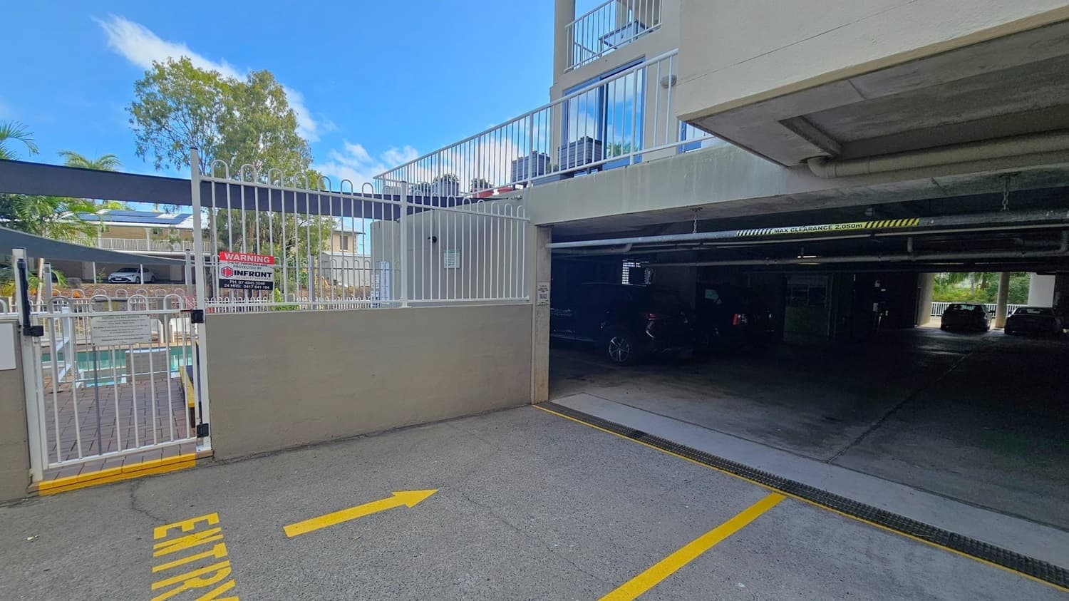 A parking area with a gated entrance on the left, featuring a white metal fence and a warning sign. Behind the gate, a swimming pool is visible. On the right, a covered parking garage with a clearance sign above the entrance. Several cars are parked inside. The ground is concrete with a yellow arrow and "ENTRY" painted on it. In the background, trees and a building are visible.