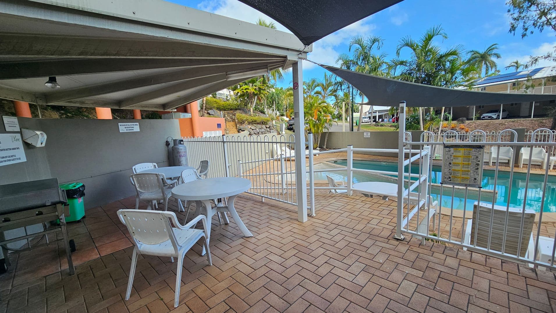 Outdoor area with a covered patio on the left featuring white plastic tables and chairs on a brick floor. A barbecue grill and a green bin are nearby. To the right, a fenced swimming pool with a few sun loungers is visible. The background includes palm trees and a building with a carport. The area is shaded by triangular fabric canopies.