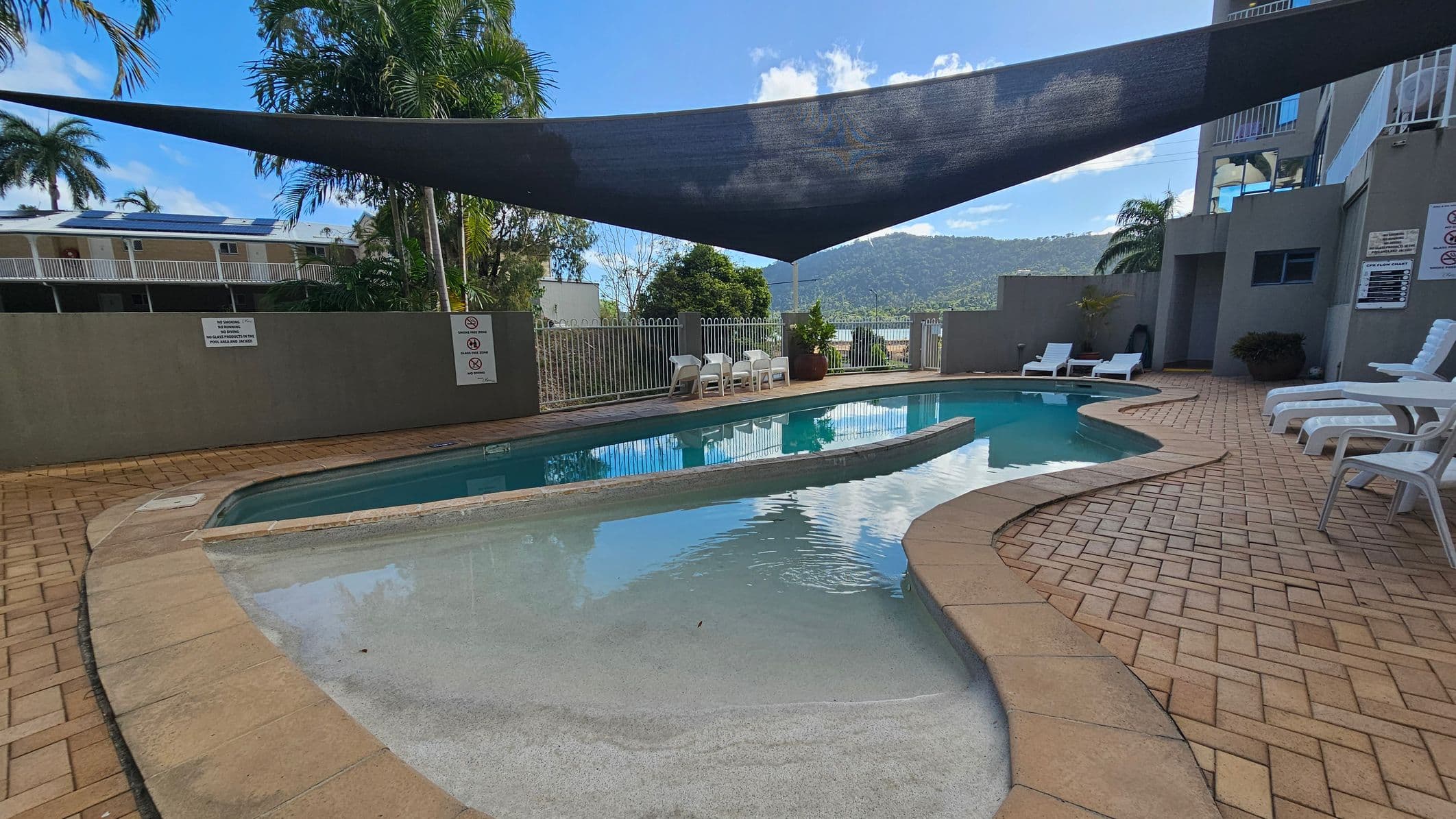 A swimming pool with a curved design is surrounded by a brick-tiled deck. On the right, several white sun loungers are arranged along the poolside. A large triangular shade sail is suspended above, providing partial cover. In the background, a grey fence with signs and a few white plastic chairs are visible. Beyond the fence, there are trees and a view of distant hills. A building with balconies is partially visible on the left.