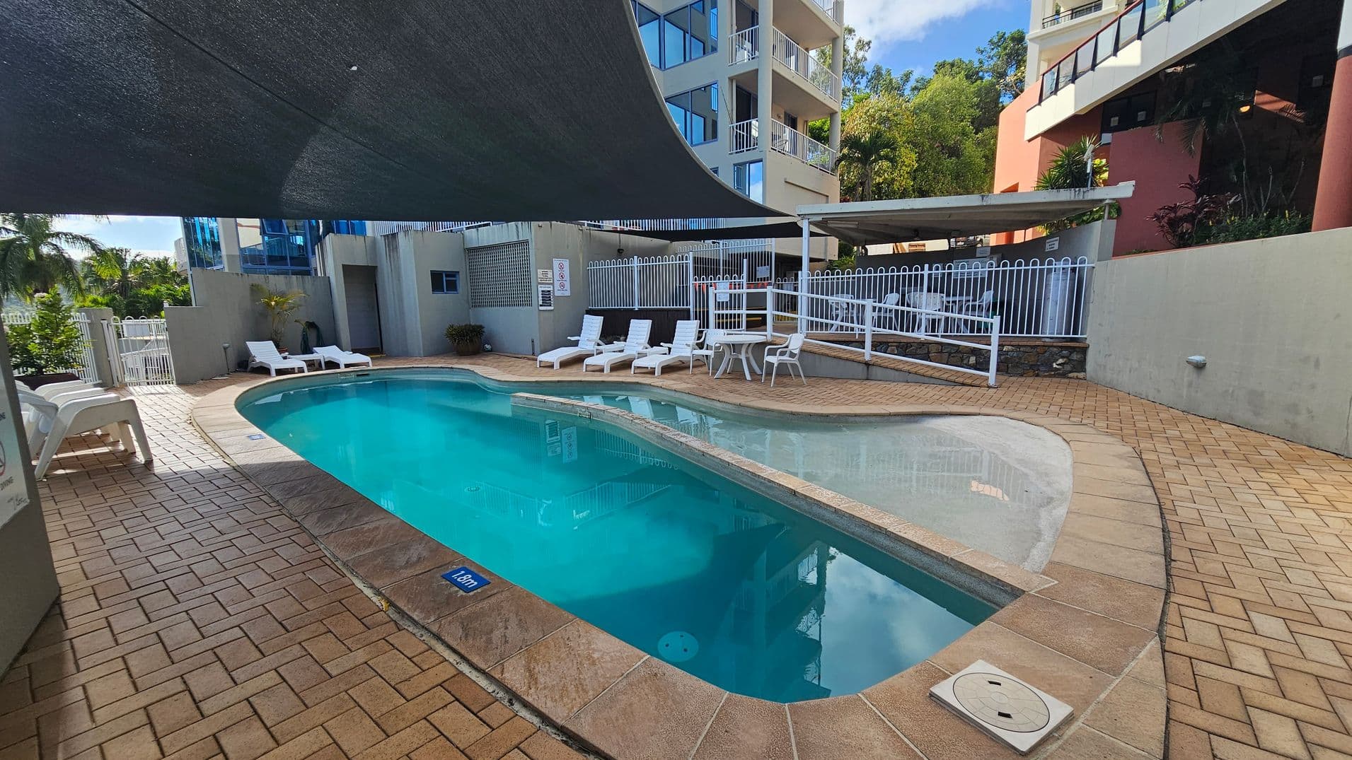 A swimming pool with clear blue water is surrounded by a brick-tiled deck. On the left, several white sun loungers are positioned under a large black shade. In the background, a multi-storey building with balconies and glass windows is visible. To the right, a white metal fence encloses the pool area, with more loungers and a small table. Lush greenery and palm trees are visible beyond the fence.