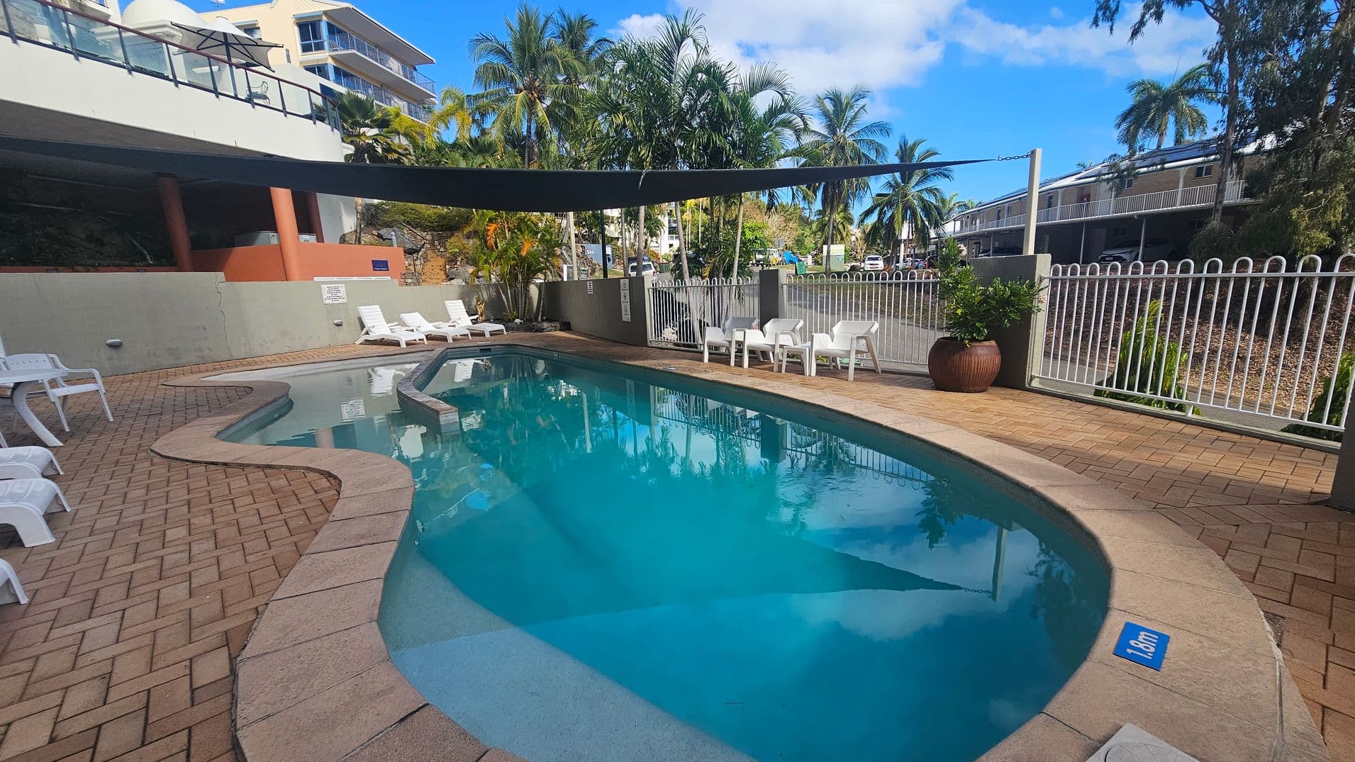 Curved swimming pool with clear blue water, surrounded by a paved deck. On the left, several white sun loungers are positioned near a low wall. On the right, a white metal fence encloses the area, with a few white plastic chairs and a large potted plant nearby. In the background, palm trees and buildings are visible, with a black shade cloth partially covering the pool area. The pool depth is marked as 1.8m.