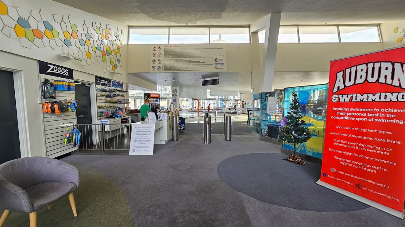 Manual inward-opening glass door at Auburn Ruth Everuss Aquatic Centre. On the upper left of the glass front wall are the conditions of entry, highlighting the prohibited activities as represented by the logos on the left. A No Smoking sign, 24/7 CCTV monitoring, and a Hiring sign are affixed to the glass doors. Information on the time when the public is limited to accessing the 25m and 50m pool is detailed on the white papers. Free entry to the facility is also detailed on the right wall.