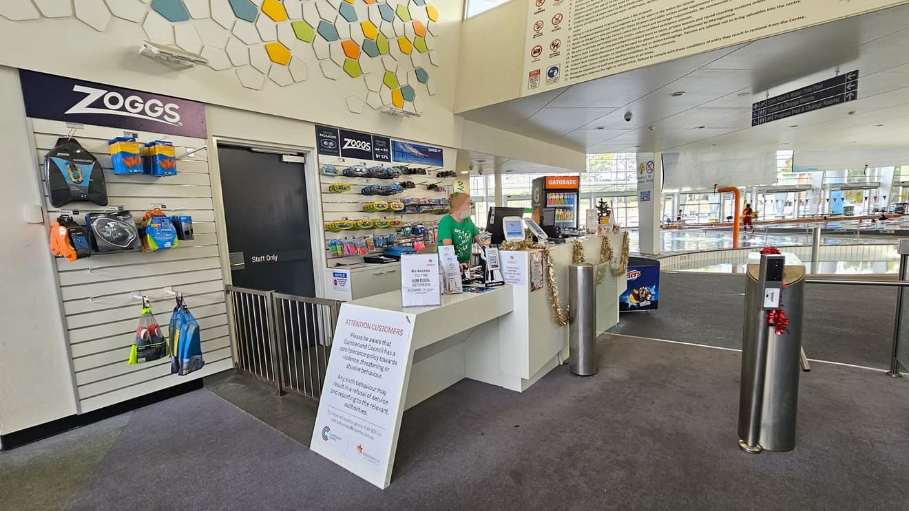 Closer view of the reception area. A small retail area that displays goggles and swimming accessories is visible behind the receptionist. Posters are placed above the low-height counter and beside the gated entrance of the reception desk. Further ahead, there is a Gatorade display placed near an ice cream freezer.
