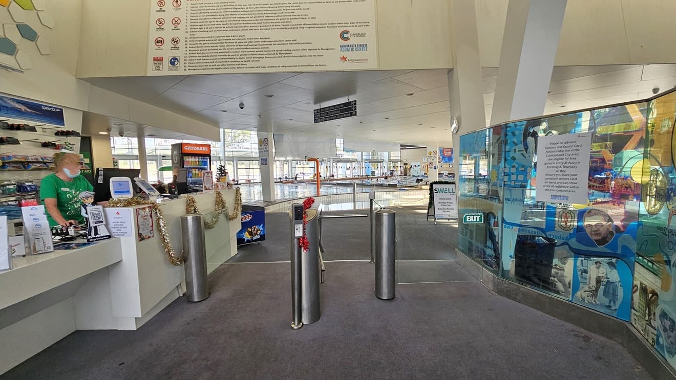 Closer view of the gated barrier leading inside the facility. The glass walls on the right side feature a collage of images and promotional materials. A signage and an exit sign are also affixed on the glass walls. A red Christmas decor is placed in the center of the gated barrier. On the left, golden Christmas decor hangs on the front of the reception desk. Further ahead, a directional sign is situated above the ceiling.