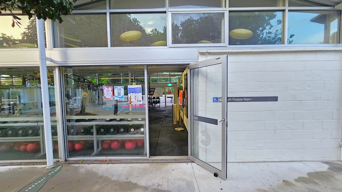 The concourse of the building is adorned with orange trees and manicured grass. The structure itself is white with glass walls. Warning and safety signs are affixed to the right wall. The non-slippery floor showcases colourful shapes in blue and red.