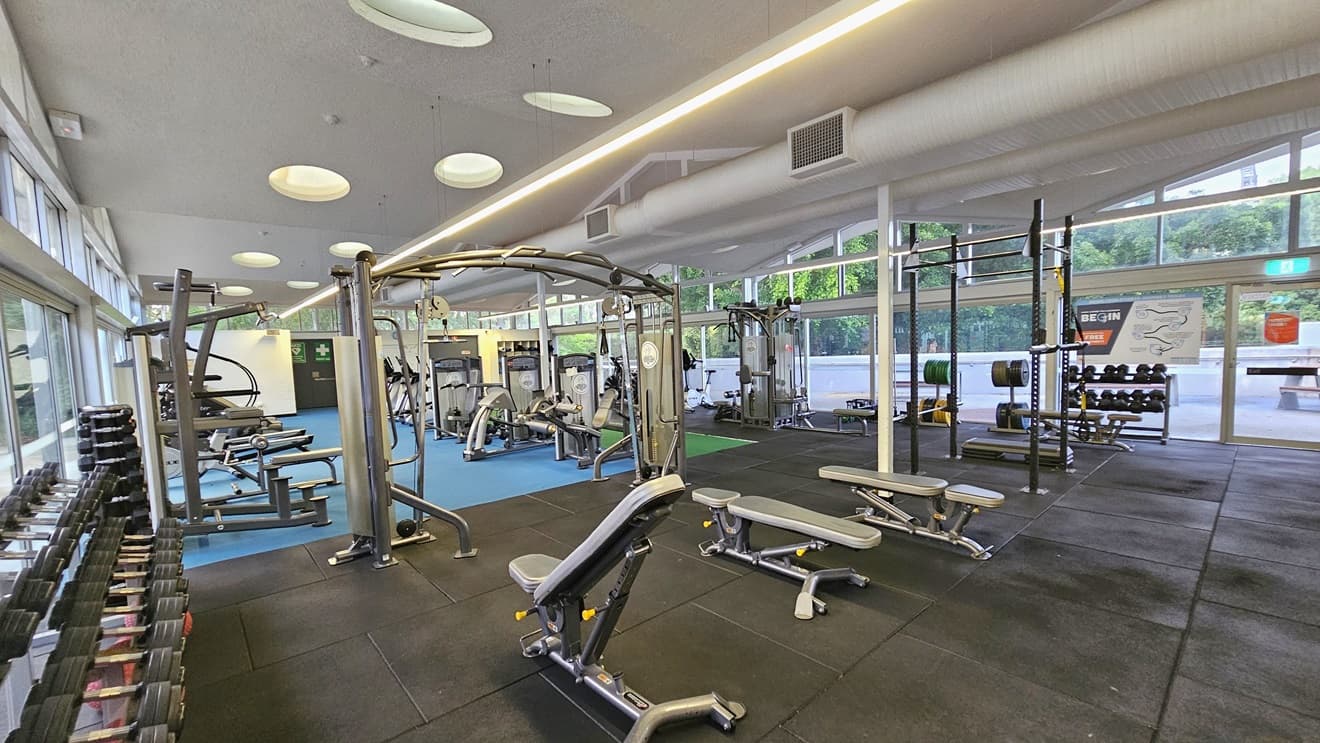 On the left, dumbbells are arranged near the wall, and lat pulldown machines are set up alongside. Bench chairs are positioned in the middle. Smith Machines and stability balls are placed to the right. Further in, more machines are arranged on the blue flooring. The weights area floor is covered with black rubber tiles. The ceiling is white, featuring LED and fluorescent lights with large aircon vents. The right wall consists of floor-to-ceiling glass with posters, offering a view outside.