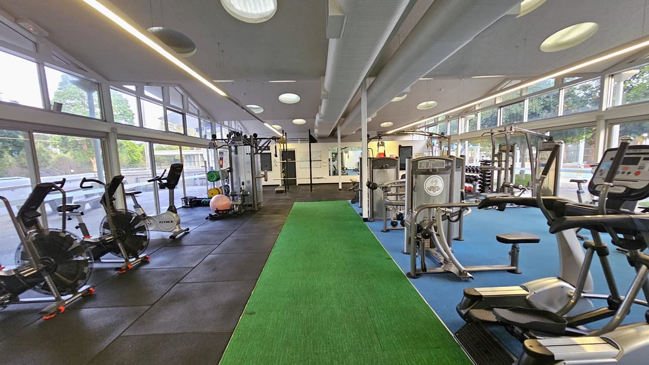 Spin machines and Smith Machines are arranged on the left, alongside lateral machines and others in the middle. Further in, free weights and Lat Pulldown machines are placed. The surrounding wall is made of glass, offering a view outside. The floor is a combination of black rubber tiles, green, and blue tiles. The ceiling is all white with LED and fluorescent lights, along with large air conditioning vents.
