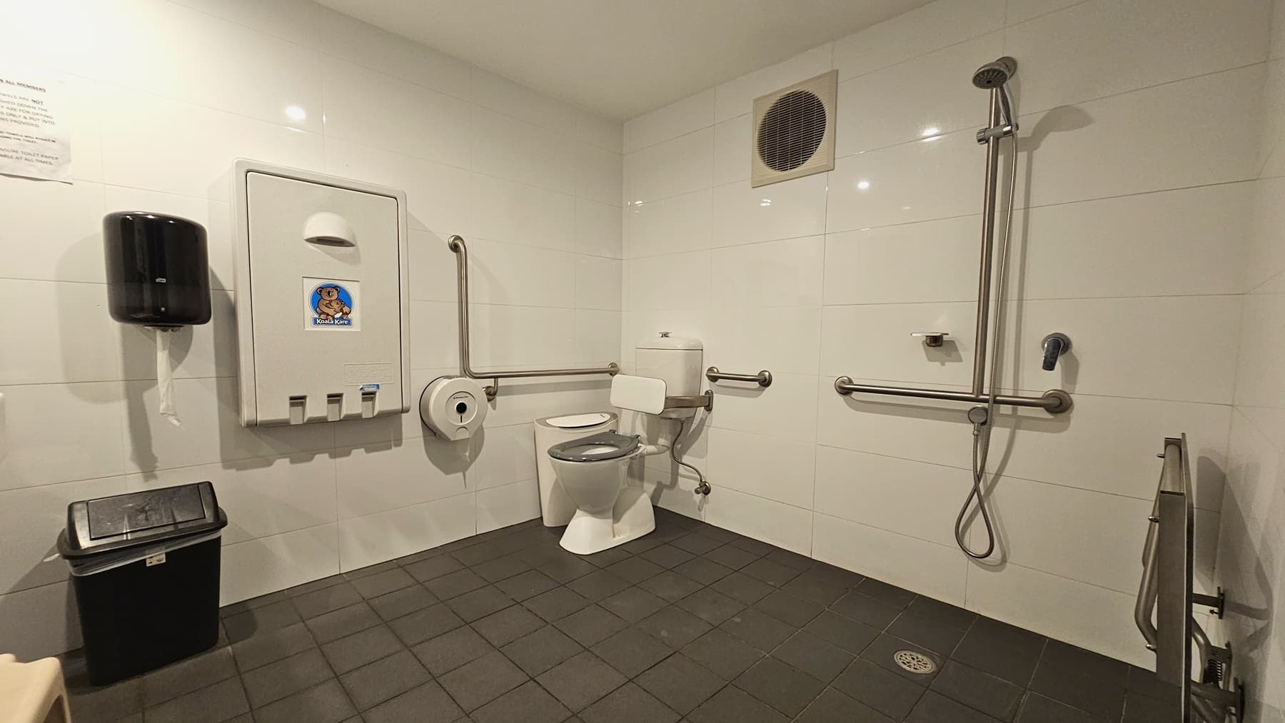 Within the accessible bathroom, a toilet bowl equipped with hand grabs is positioned on the left, accompanied by a rubbish bin. On the left wall, there's a tissue dispenser, a foldable change table, and another rubbish bin. To the right, a shower with handrails is present, and a foldable bench is positioned against the wall. The floor is covered with grey tiles, and the walls have an exhaust fan, while the ceiling is white.
