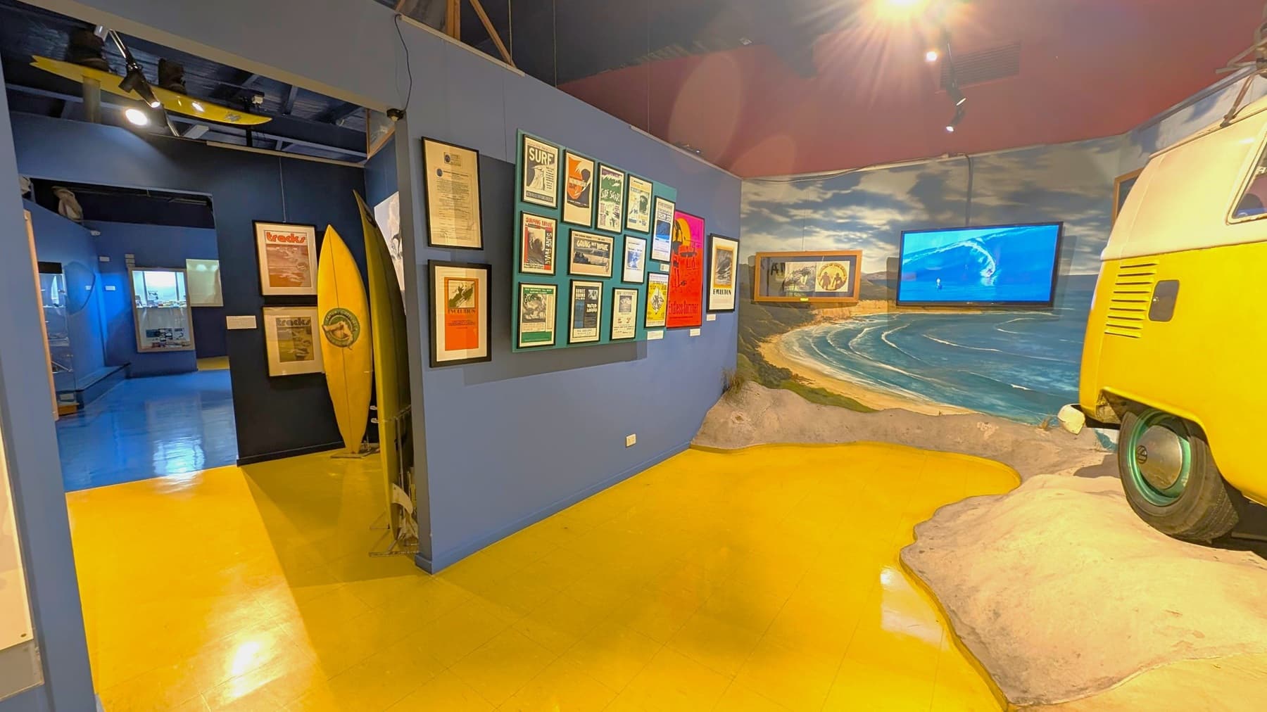 A museum exhibit with a surf theme. On the left, there are framed posters and a yellow surfboard against a dark wall. The floor is yellow. In the middle, a wall displays various framed surf-related posters. On the right, a mural of a beach scene with a screen showing a surfing video. The front of a yellow van is partially visible, positioned on a sandy display. The ceiling is dark with spotlights.