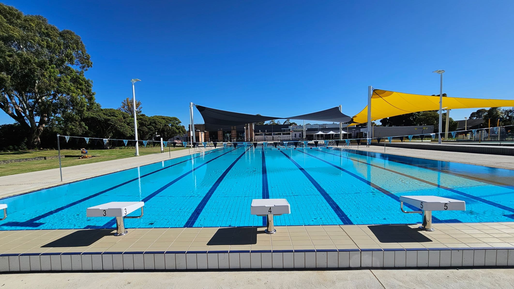 An outdoor swimming pool with clear blue water and marked lanes. Starting blocks numbered 3, 4, and 5 are visible at the pool's edge. On the left, there are large trees and a grassy area. On the right, there are yellow and black shade sails above the poolside, with more trees and a paved area. In the background, there are buildings and additional seating areas.