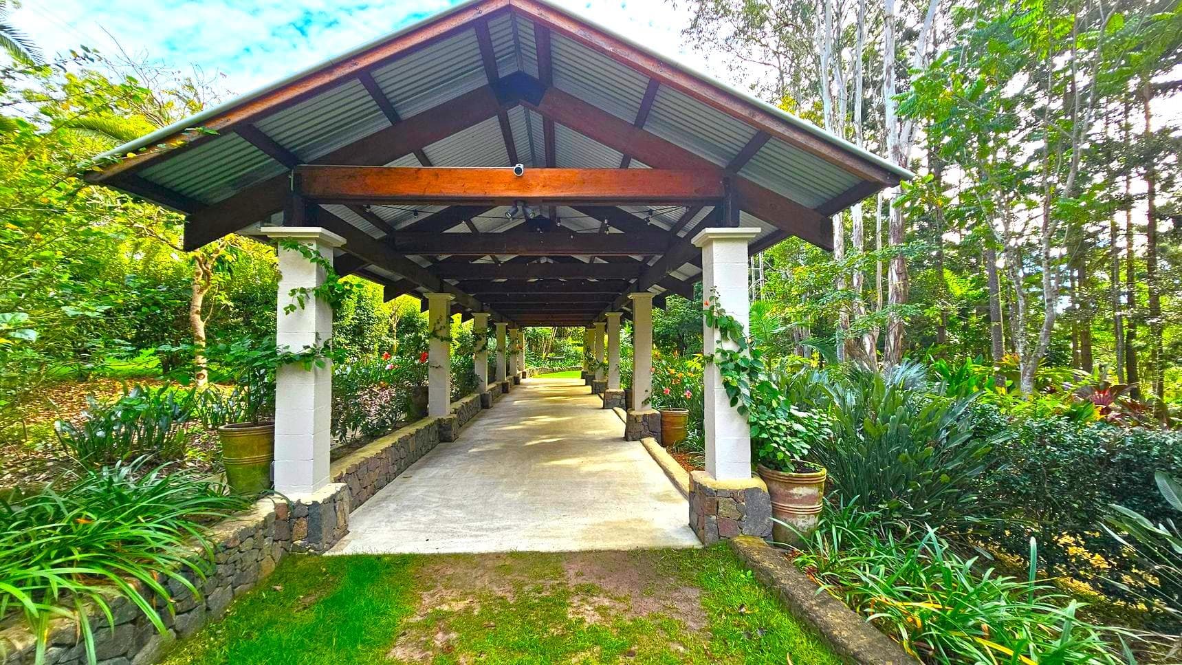 A covered walkway with a metal roof supported by stone pillars extends into the distance. The path is flanked by lush greenery and potted plants on both sides. The ground is a mix of grass and concrete, and the surrounding area is filled with tall trees and dense foliage, creating a serene, natural environment.